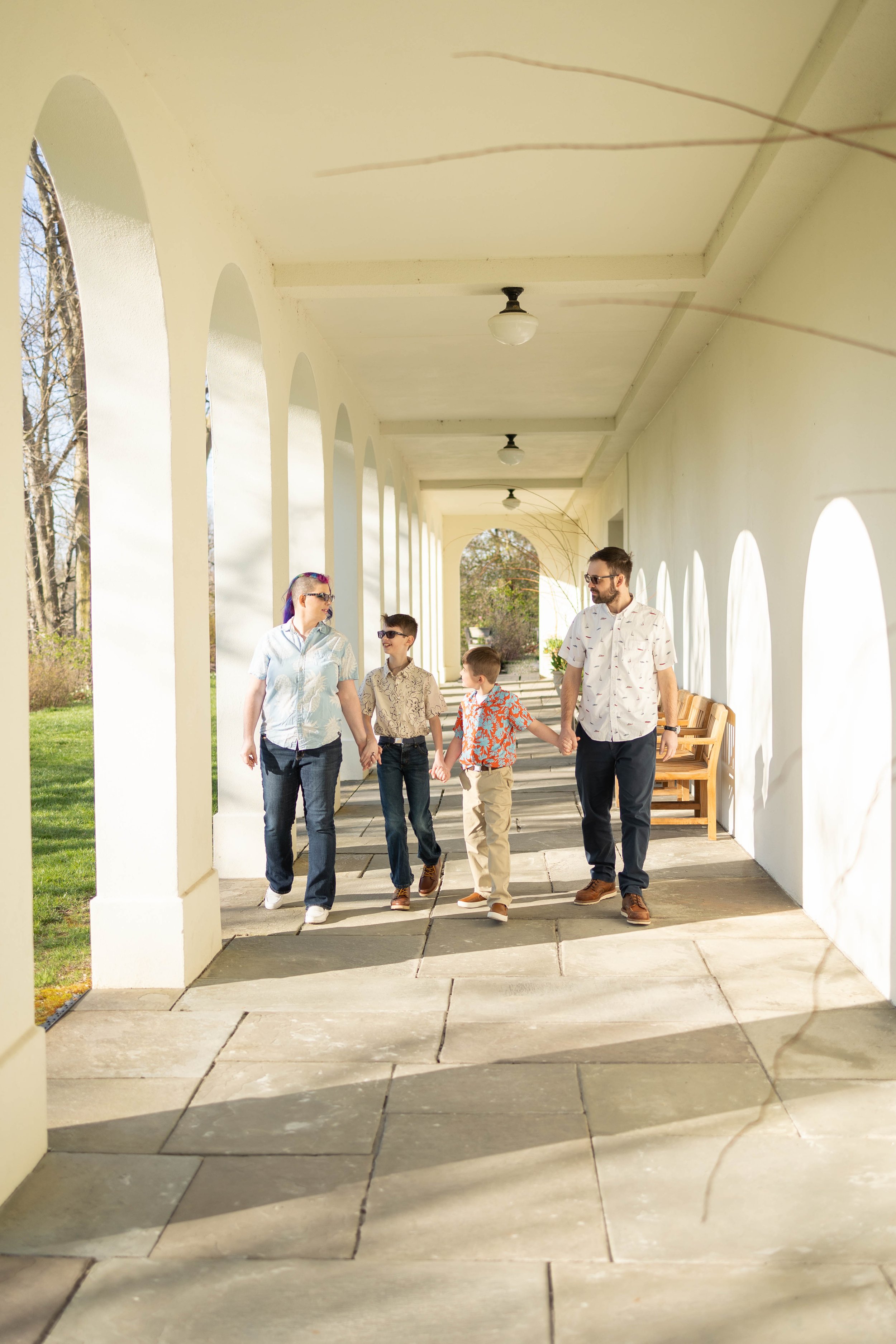 A family of four holding hands, walking through a covered porch on a sunny day.