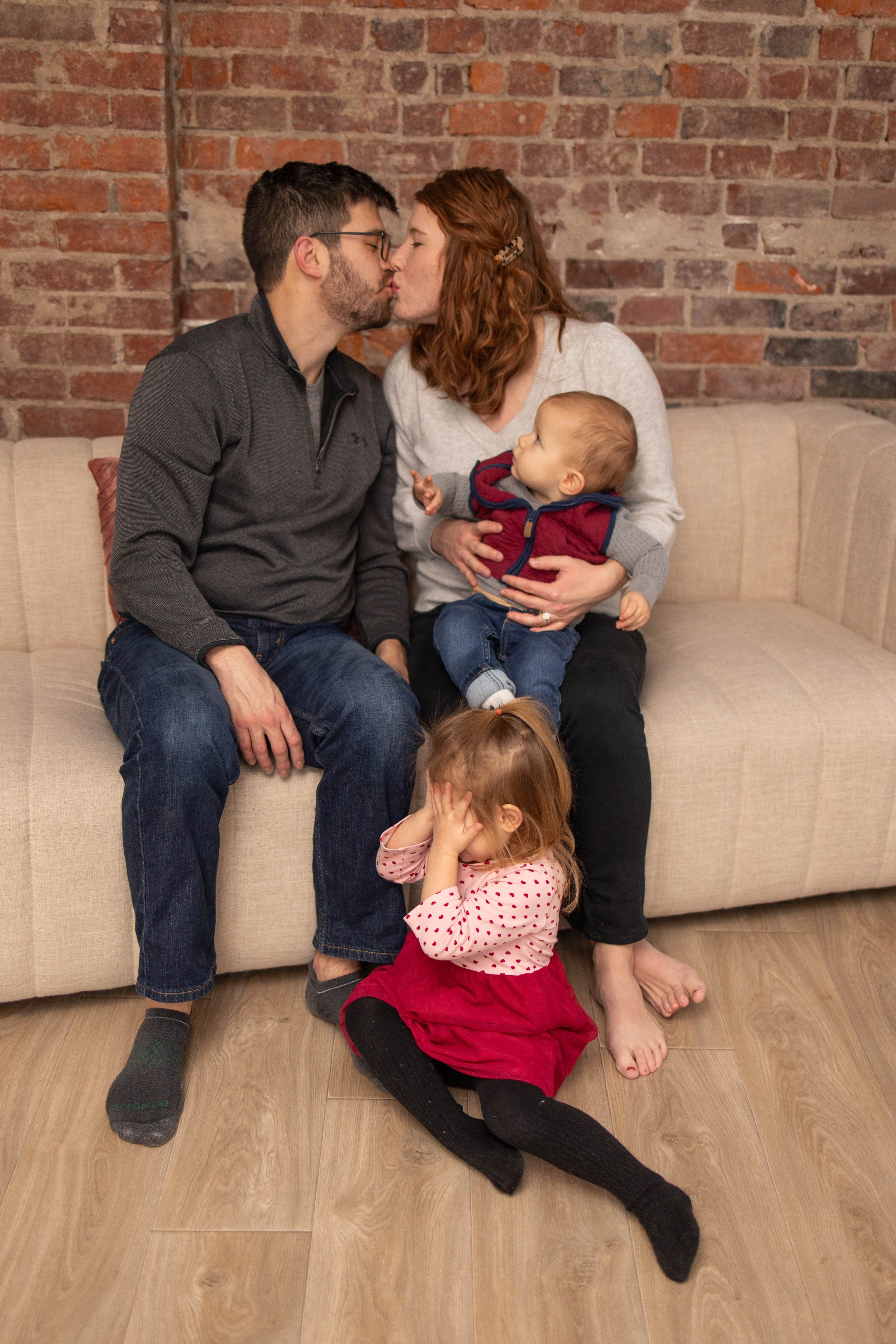 A family of four sitting on a beige couch in front of a brick wall. The couple is kissing, with the mother holding a young boy. A little girl is on the floor covering her ears.