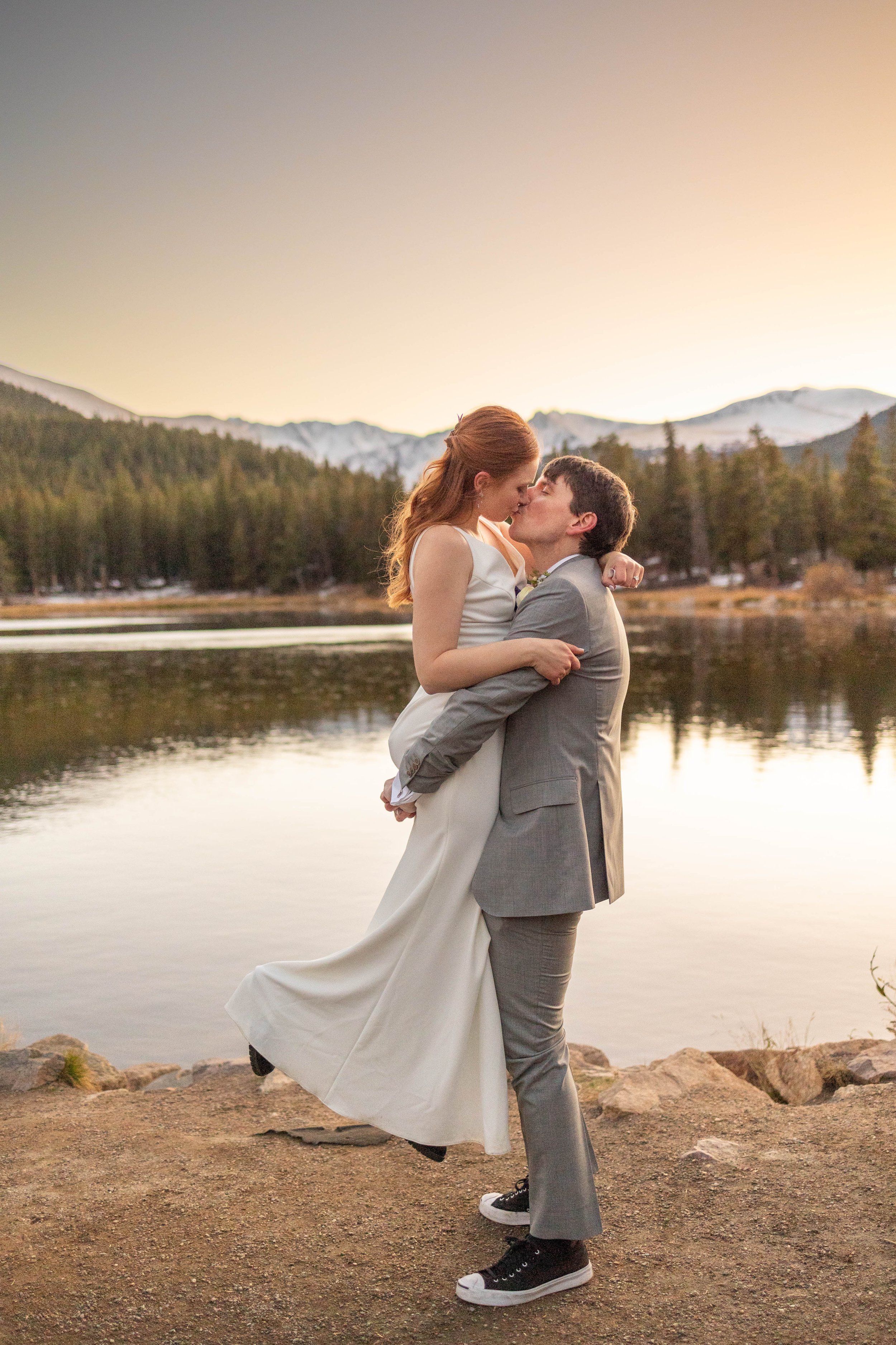 A couple kissing by a lakeside at sunset, with mountains and trees in the background.