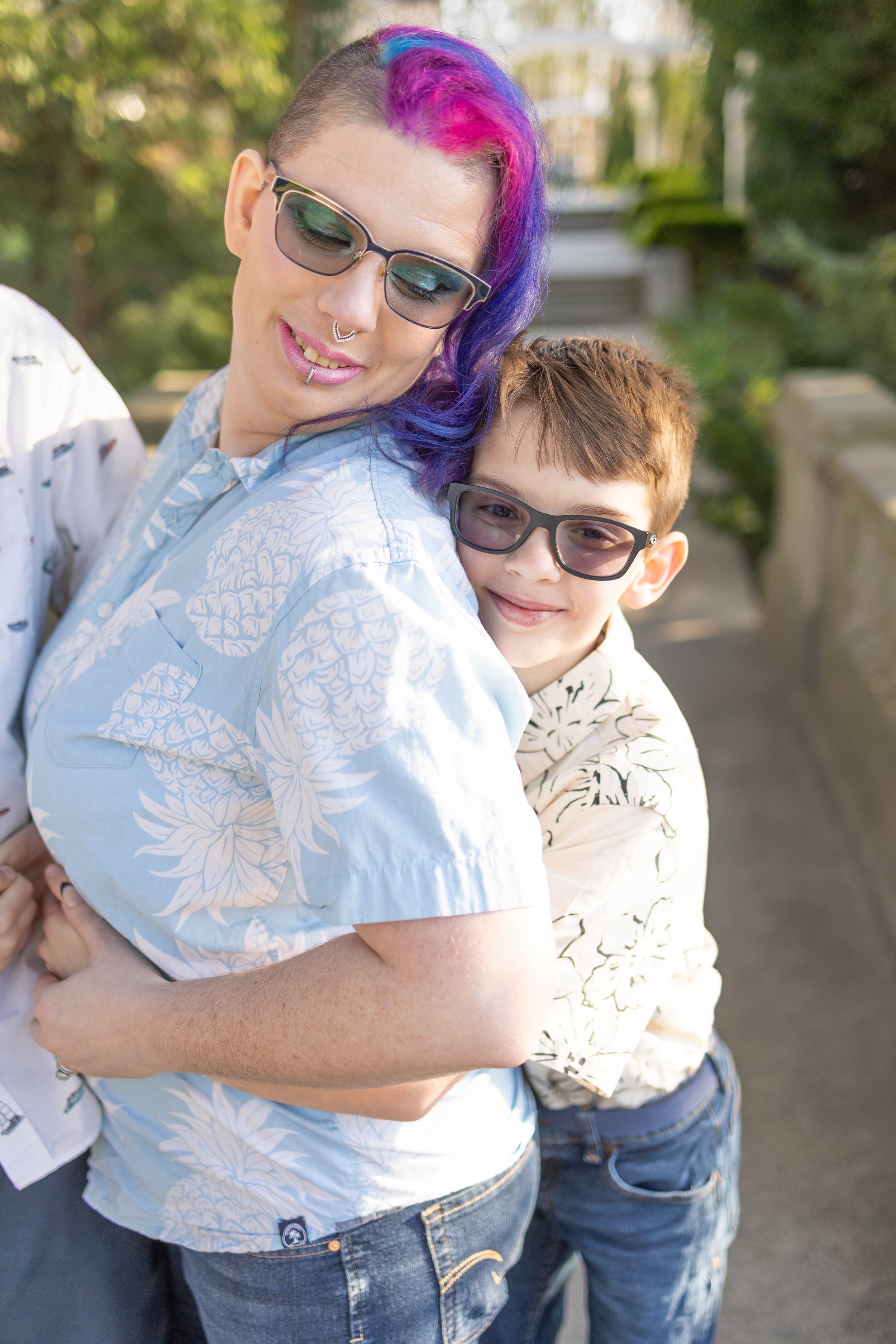 A woman with colorful purple and pink hair and a young boy with short brown hair are hugging each other outdoors. Both wearing sunglasses and smiling.