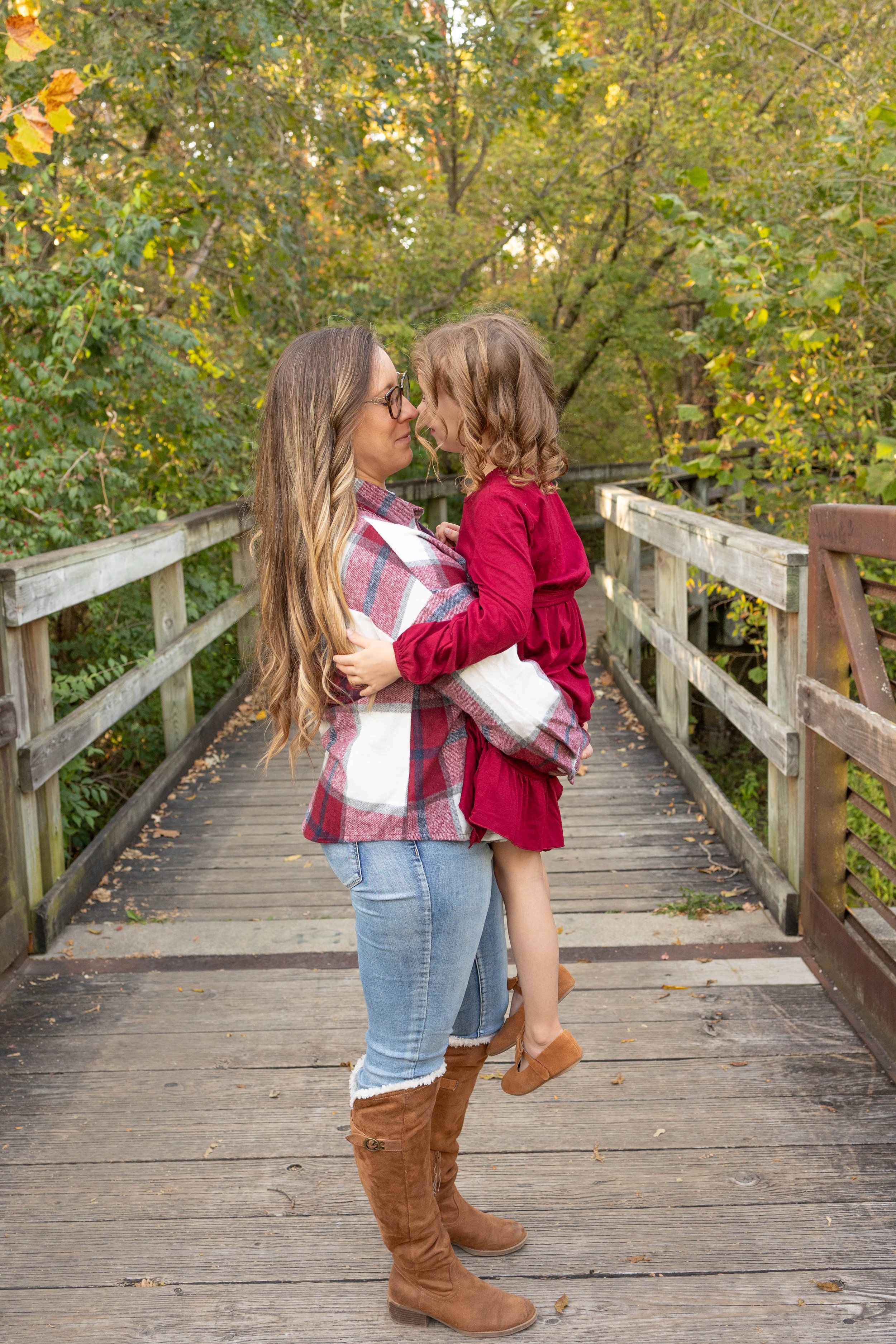 A woman holding a young girl in her arms on a wooden bridge surrounded by autumn foliage.
