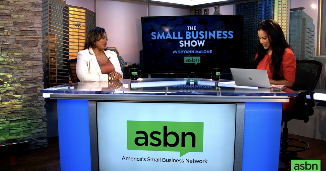 Two women sitting at a news desk with a large screen behind them displaying the text "The Small Business Show w/ Shyann Malone". The desk has an 'asbn' logo with the tagline "America's Small Business Network". One woman is dressed in a white blazer and the other in a red dress, with city buildings visible through the windows in the background.