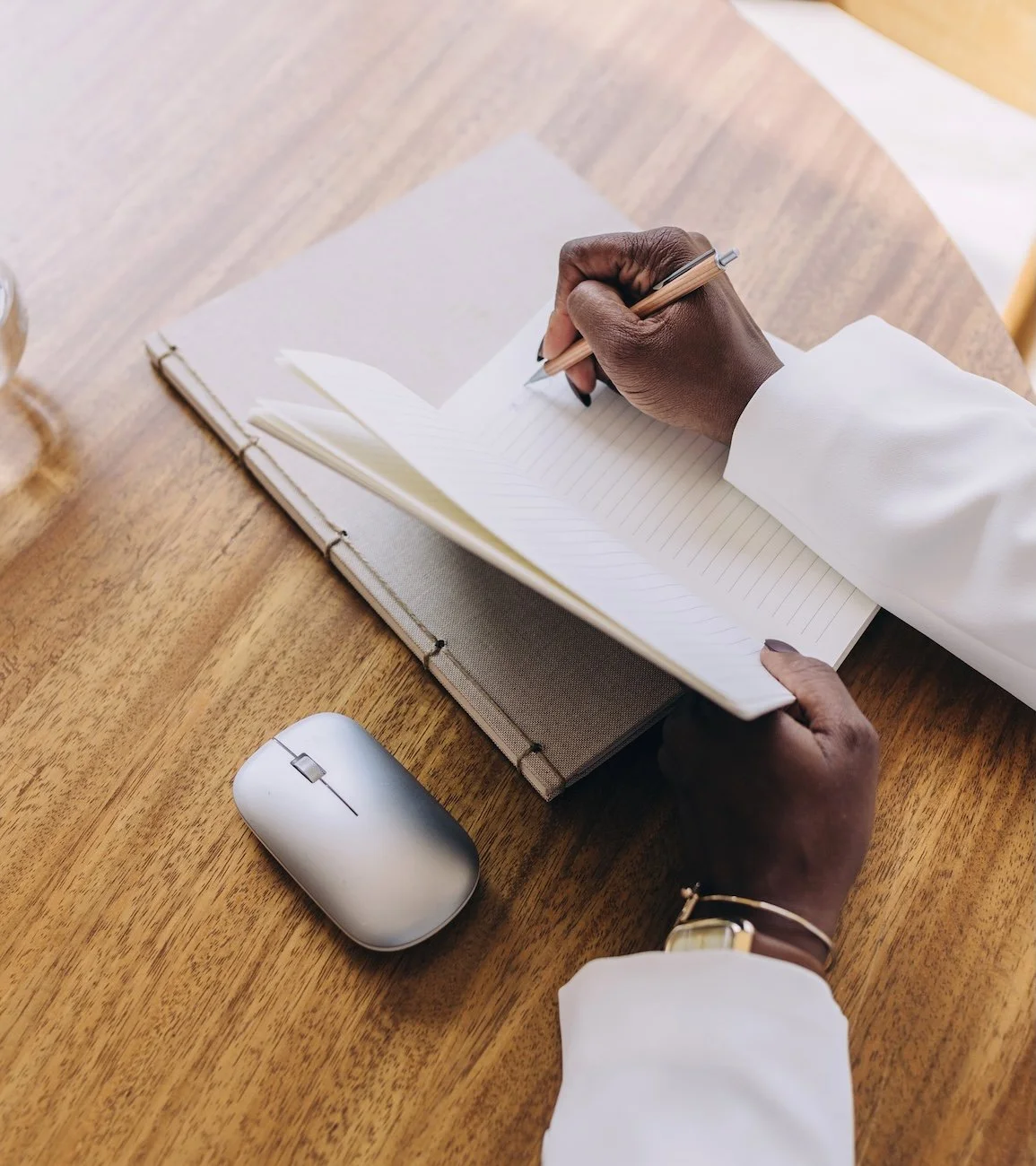 A person writing in a notebook at a wooden desk with a computer mouse nearby, wearing a white shirt and a bracelet.