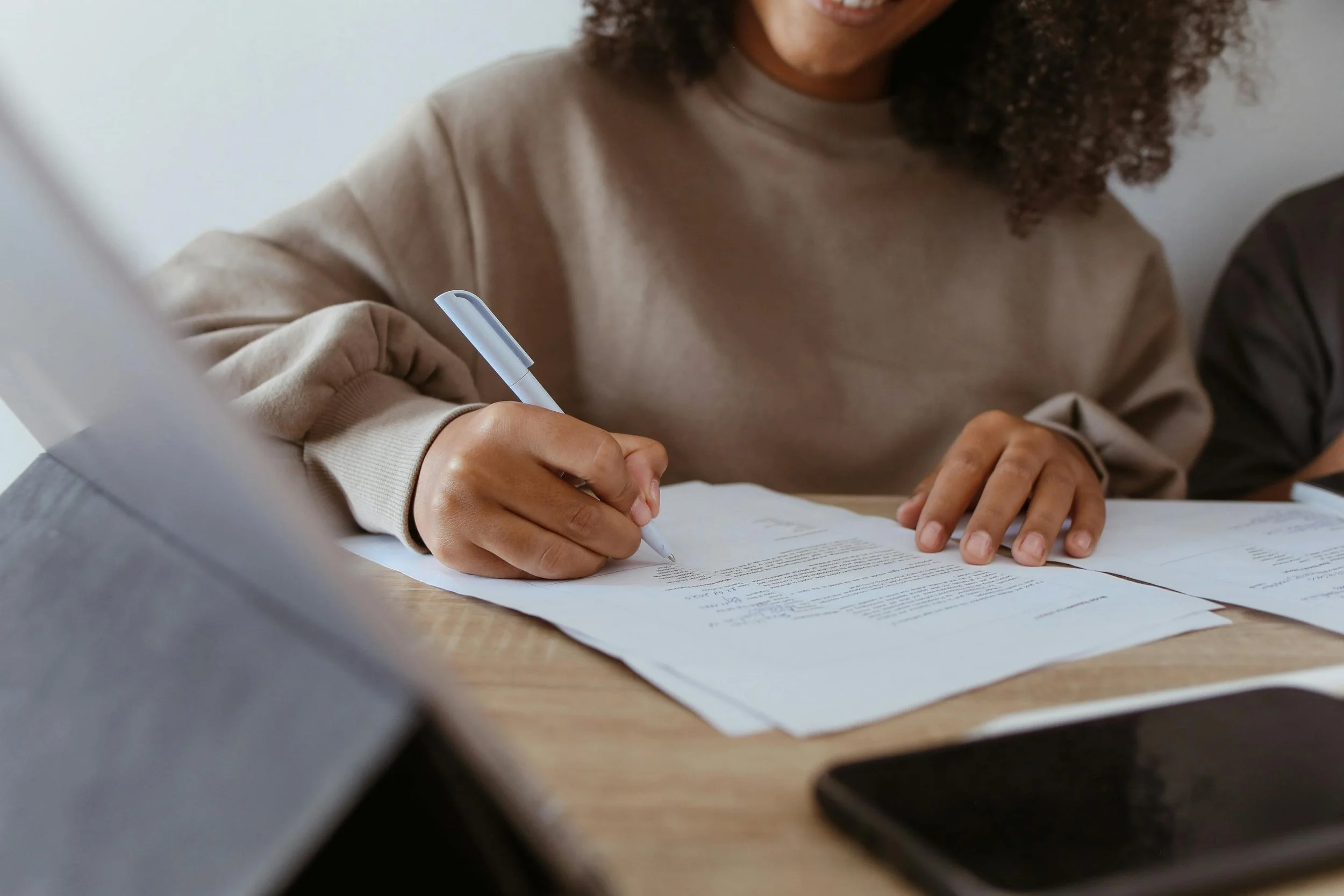 Person writing on a paper with a pen on a wooden desk, with a smartphone and additional papers visible.