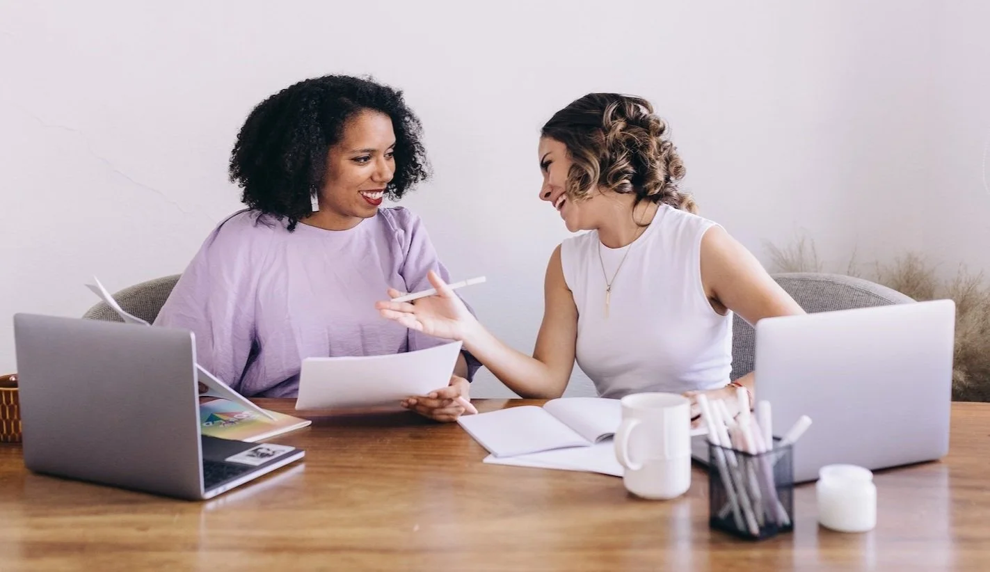 Two women working at a desk, smiling and talking, with laptops, notebooks, and pens.