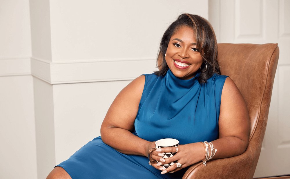 A woman with dark hair and a bright smile sitting in a brown chair, wearing a sleeveless blue dress, holding a white mug with a black geometric pattern.