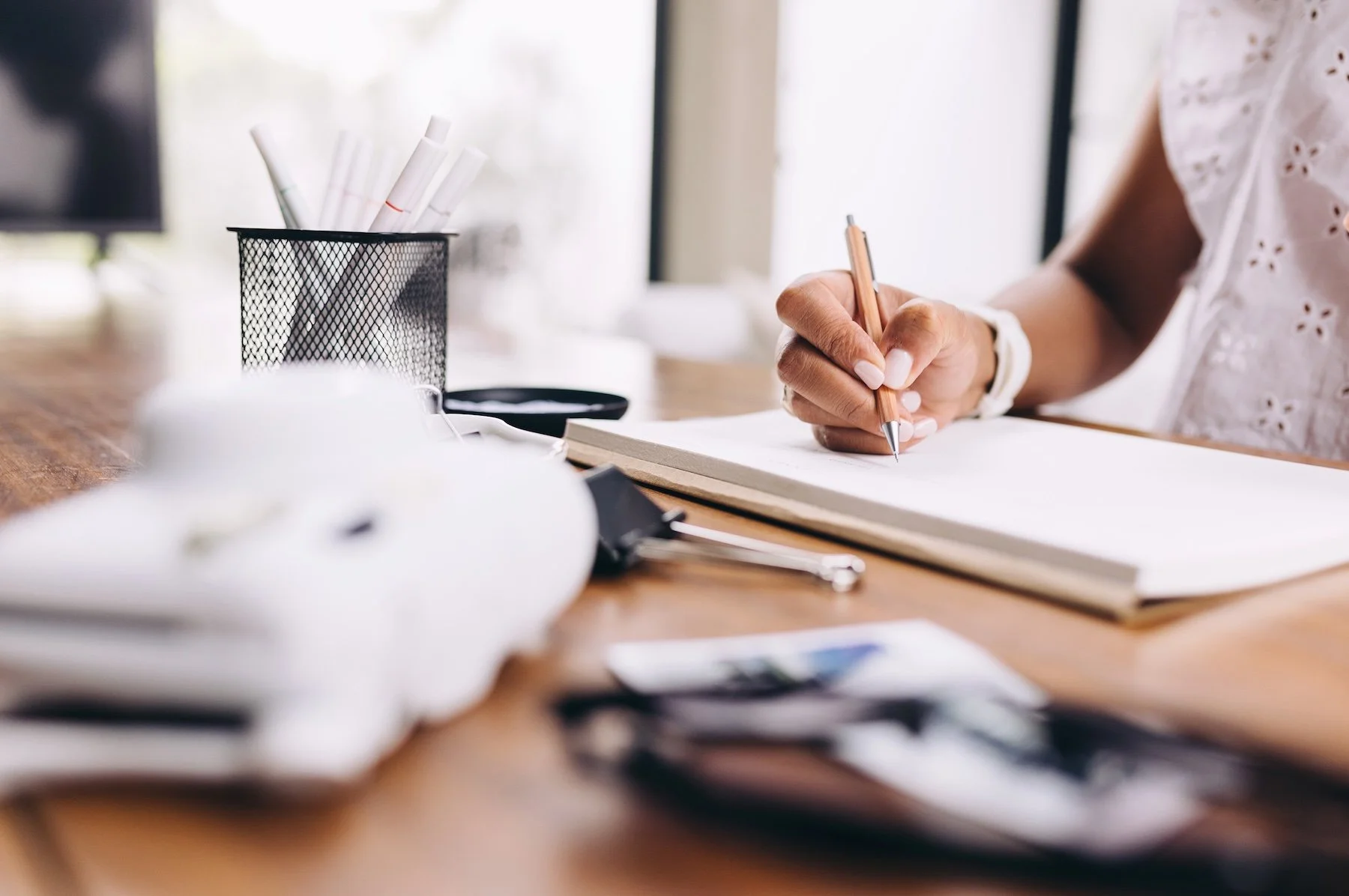 A person writing in a notebook on a wooden desk with office supplies and blurred papers around.