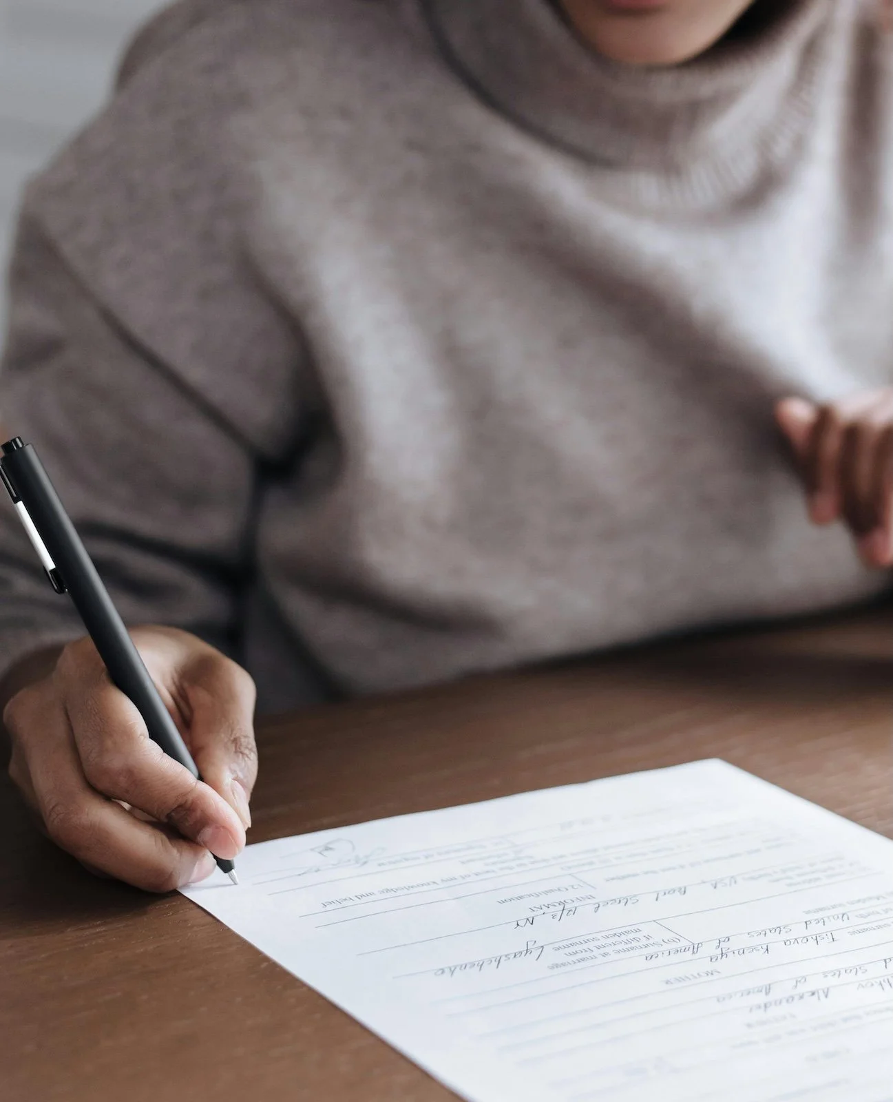 A person in a gray sweater is writing on a form with a black pen at a wooden table.