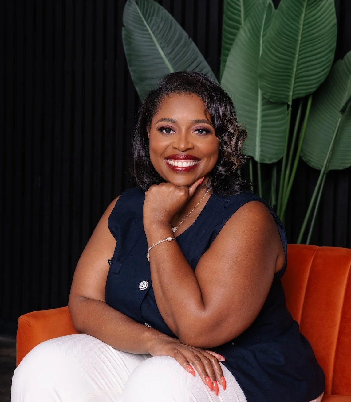 A smiling Black woman with shoulder-length wavy hair sitting on an orange chair with large green leaves in the background, wearing a navy sleeveless top and white pants.