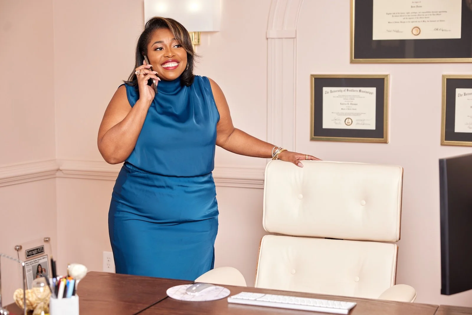 A woman in a blue dress smiling and talking on a phone in an office setting.