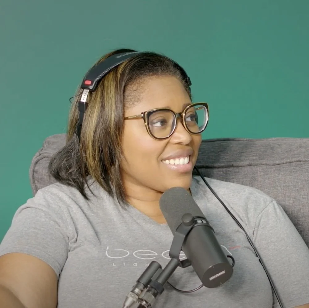 A woman with glasses, headphones, and a gray t-shirt sits in front of a microphone, smiling and looking to her right.