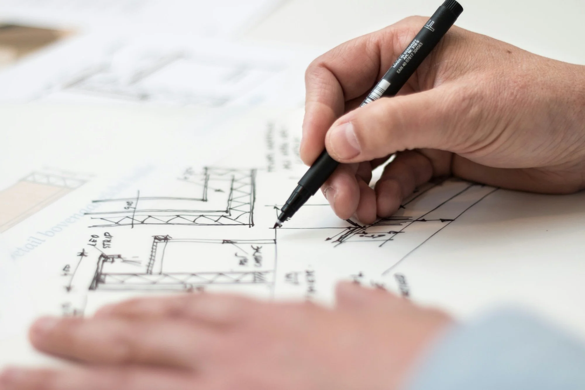 A person's hand sketches architectural drawings with a black marker on paper, showing structural design and measurements.