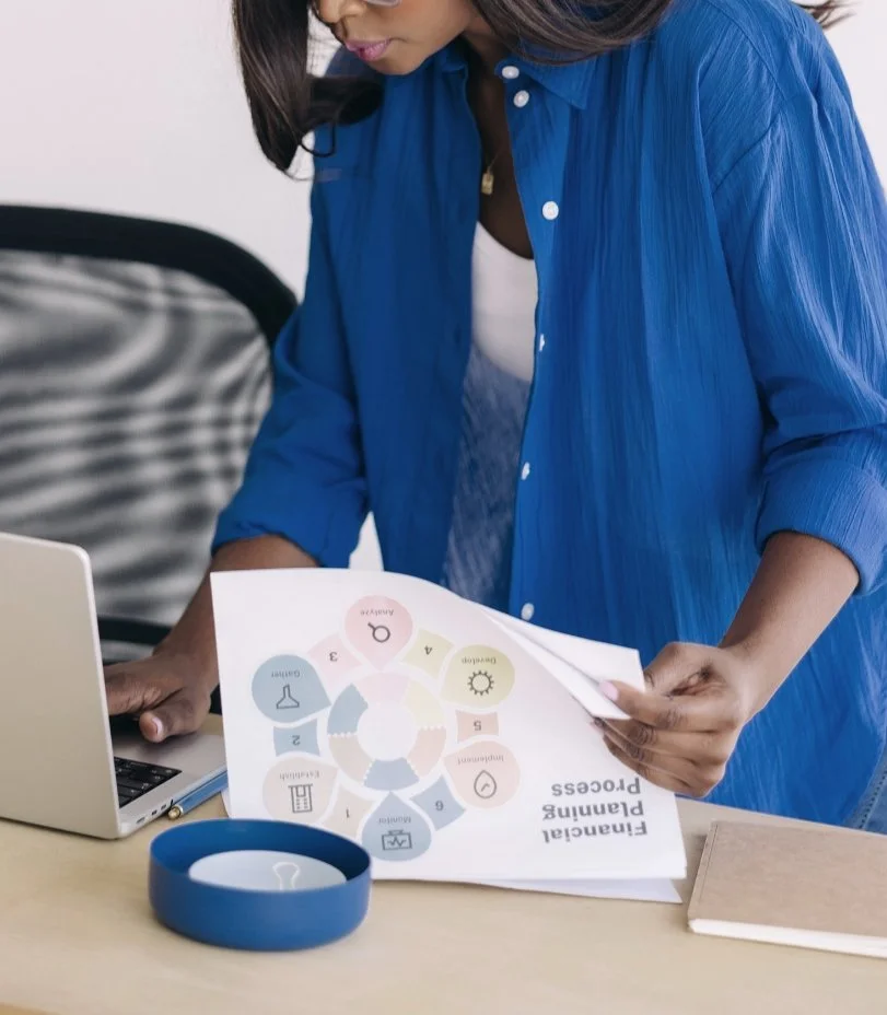 A woman in a blue shirt looking at a printed planogram with colorful icons and text, sitting at a desk with a laptop and a blue container.