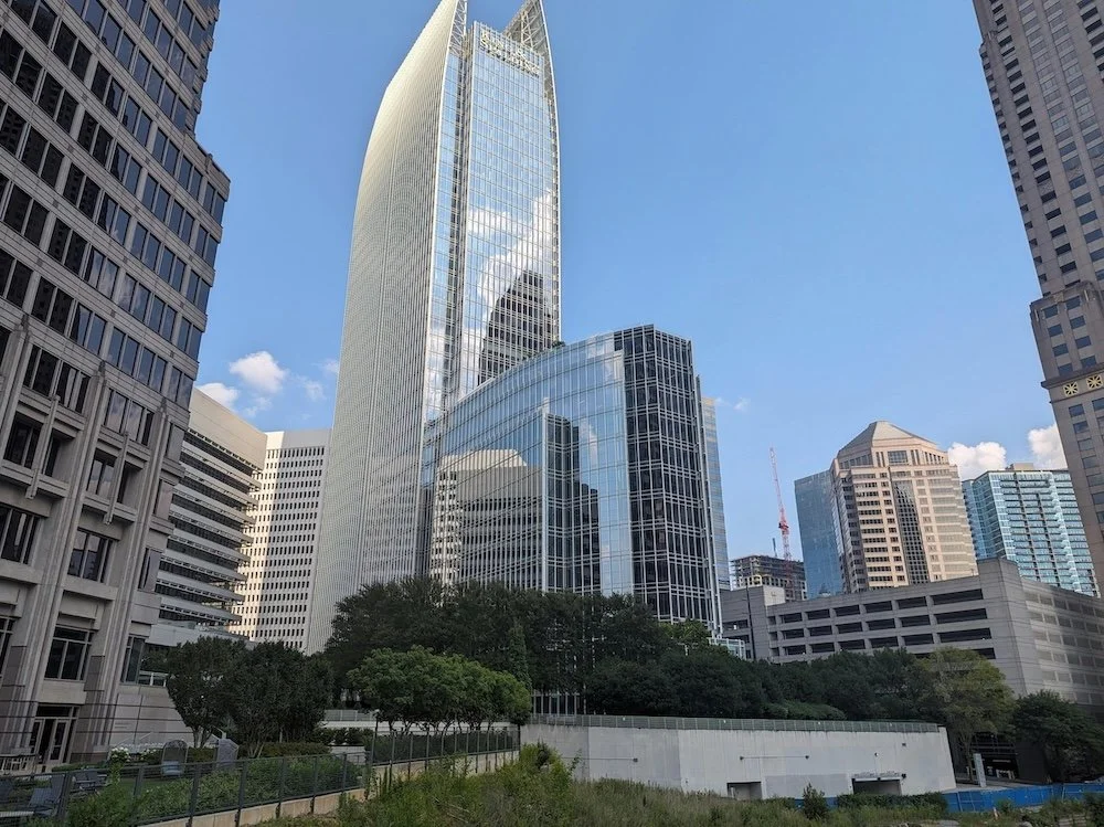 Tall modern skyscrapers with glass facades in a city skyline under a blue sky, with trees and a park area in the foreground.