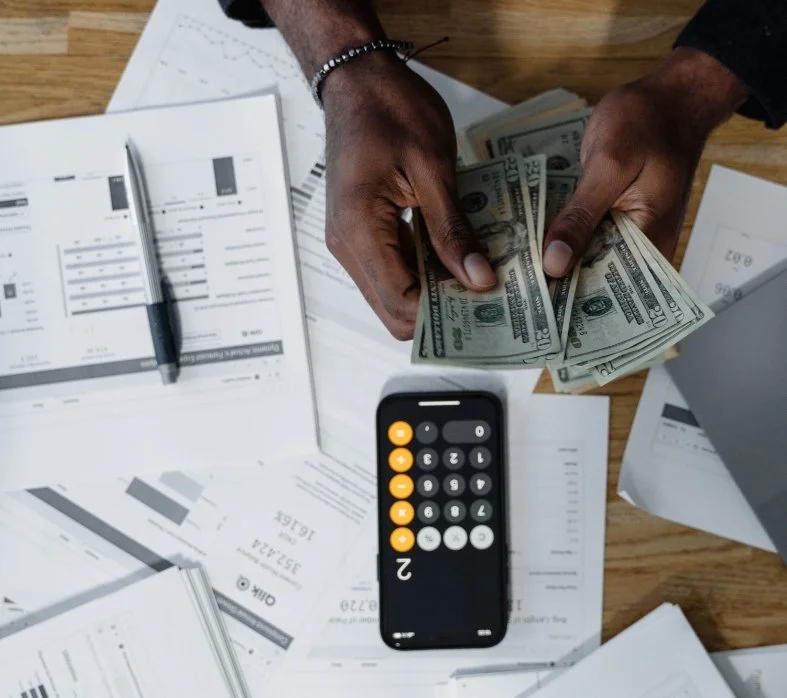 Hands counting cash amidst financial documents, a calculator, and a pen on a wooden desk.