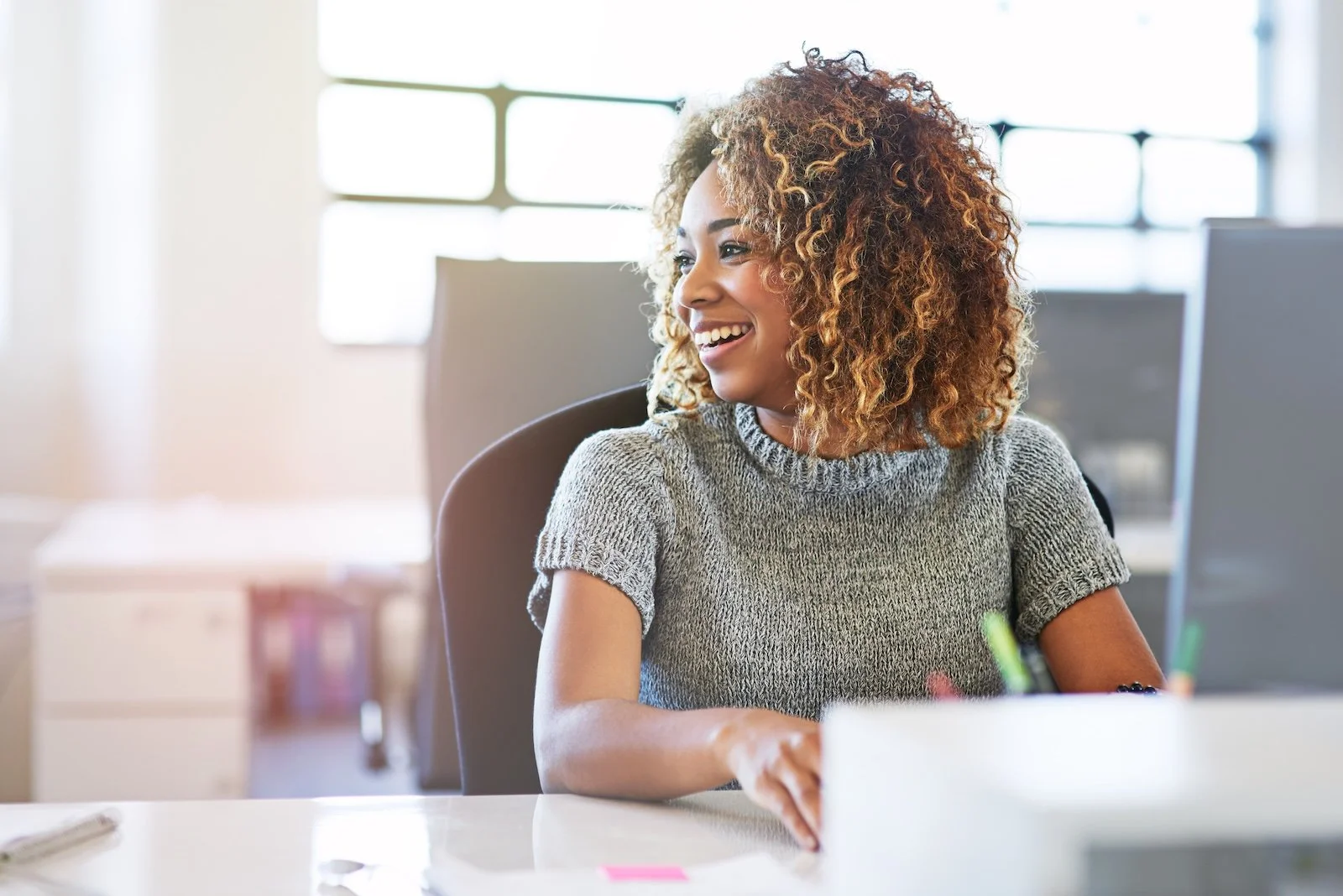 A smiling woman with curly hair working at her desk in an office setting.