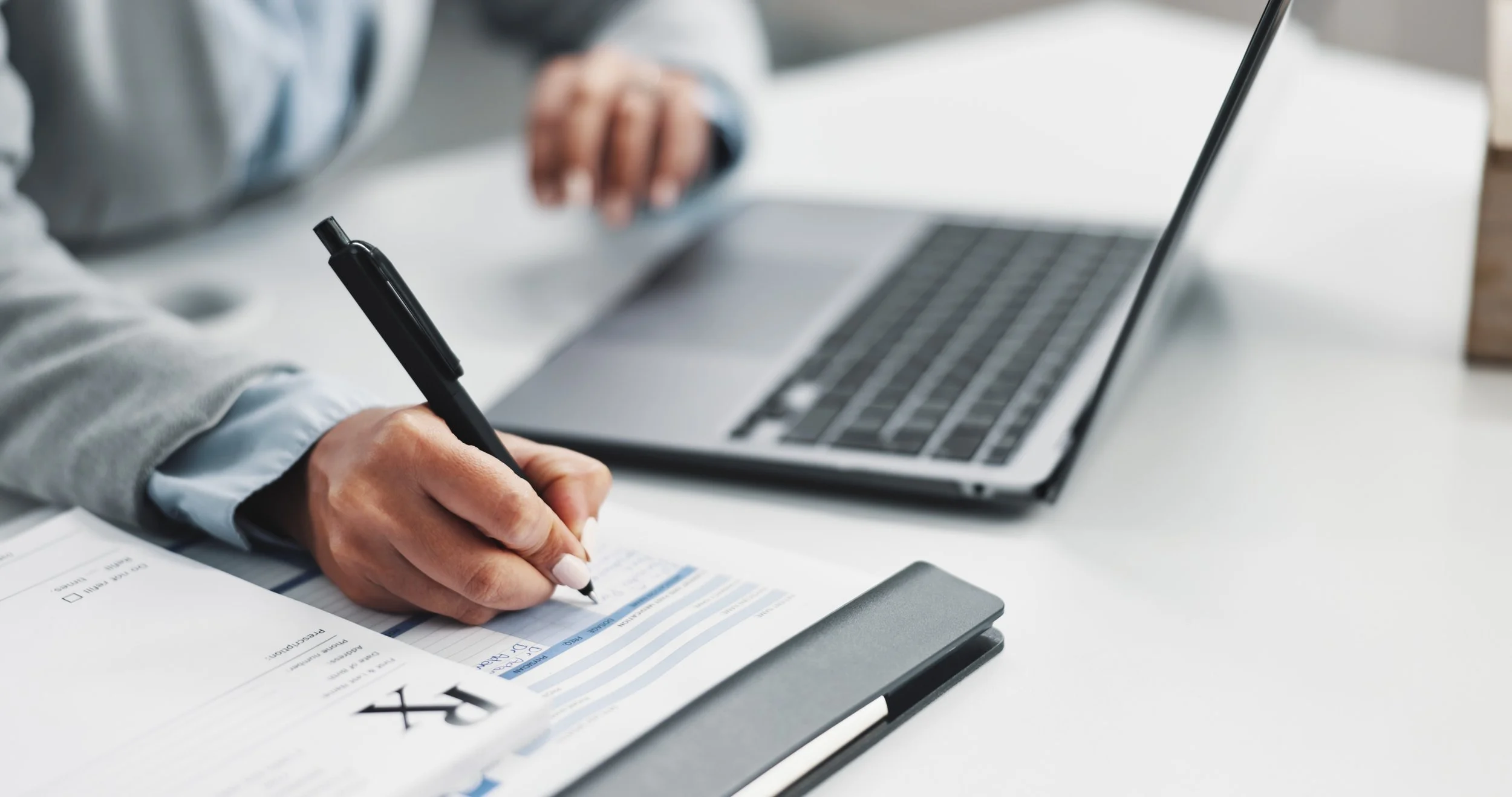 A person writing on a financial report or document with a pen, with a laptop open on a white desk.