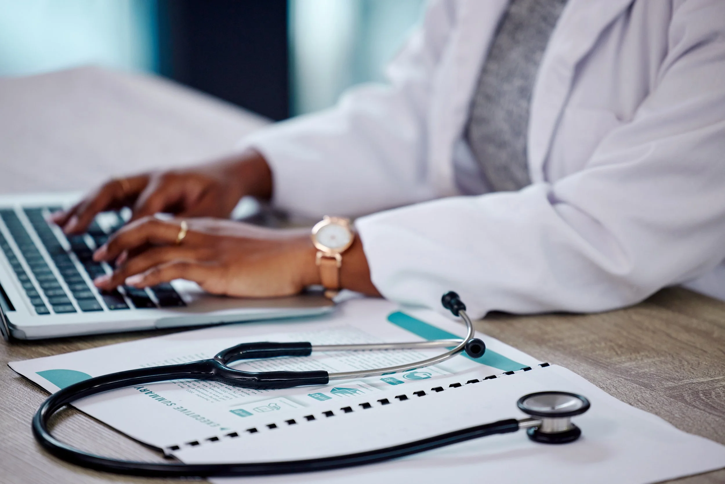 A person in a white doctor's coat typing on a laptop with a stethoscope and medical documents on the table.