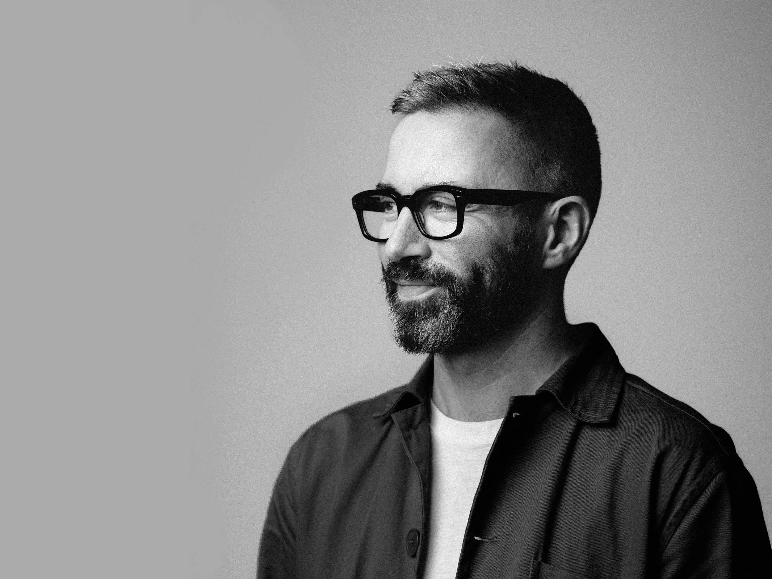 Black and white portrait of a man with glasses, beard, and short hair, looking to the right, wearing a dark jacket over a light shirt.