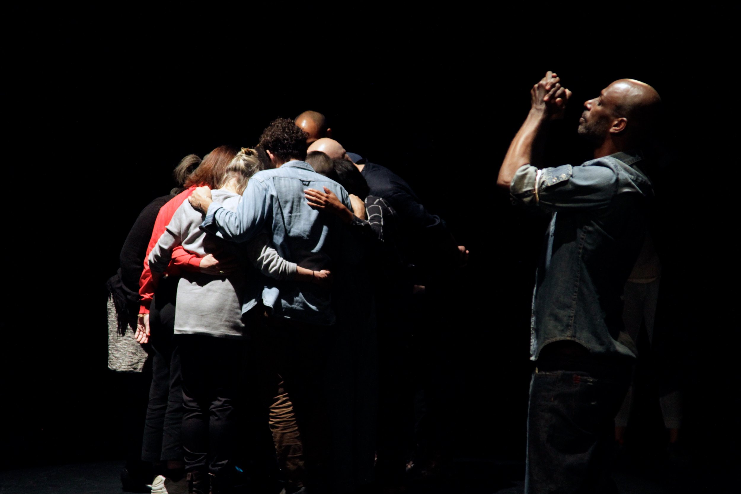 A performer stands with hands clasped in front of his face near a group huddled with arms around each other. Photo by Simon Courchel.