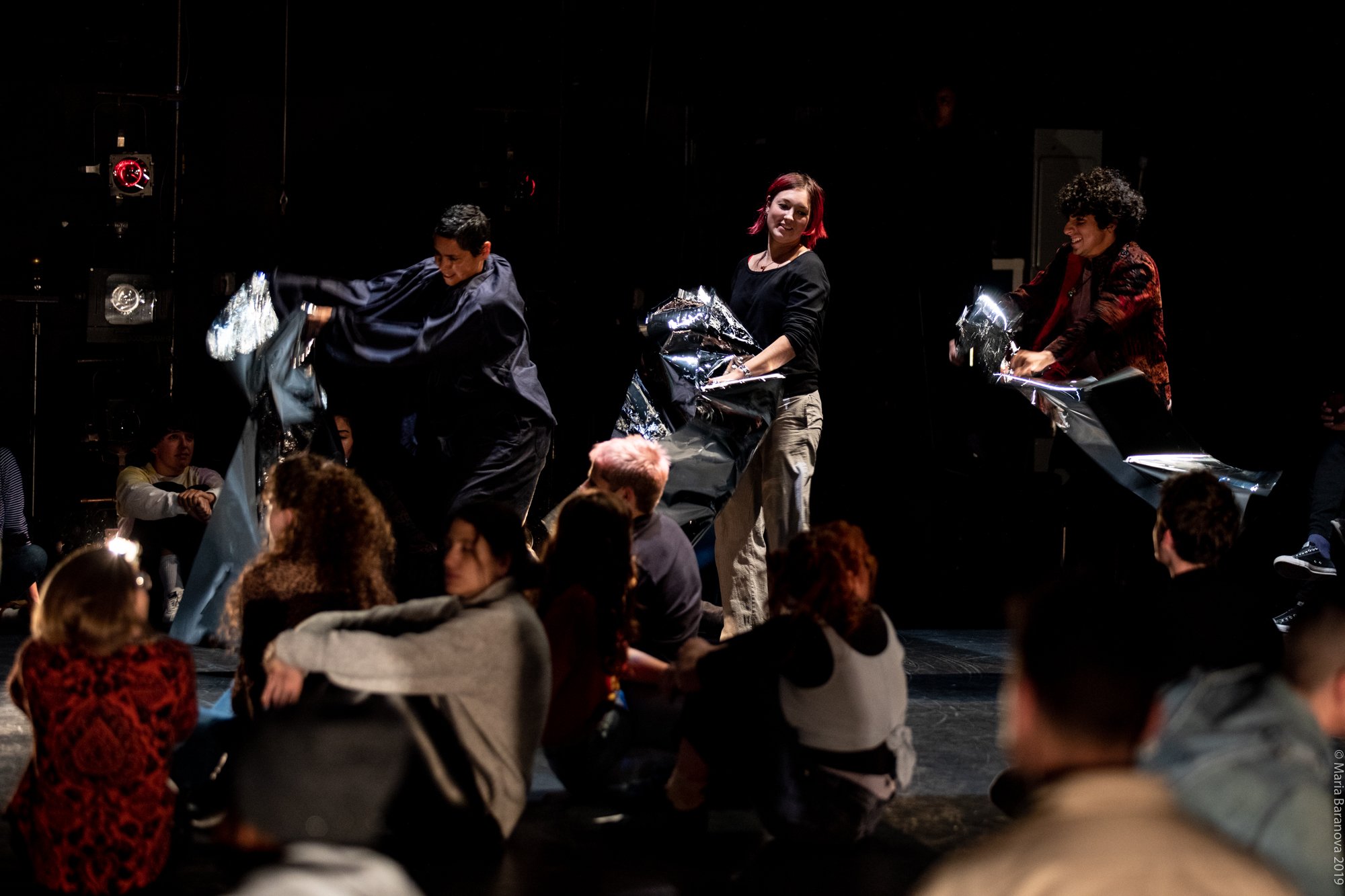 Three people wave sheets of mylar on a stage while a group sits in front of them. Photo by Maria Baranova.