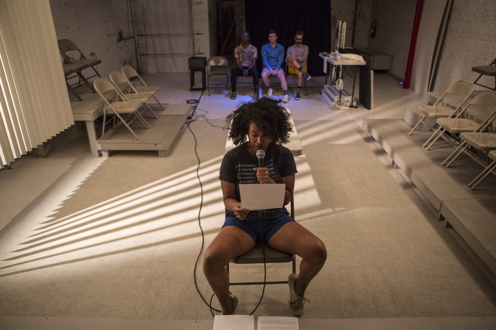 A performer sits reading from a page into a microphone. Three other performers look on from behind. Photo by Maria Baranova. 