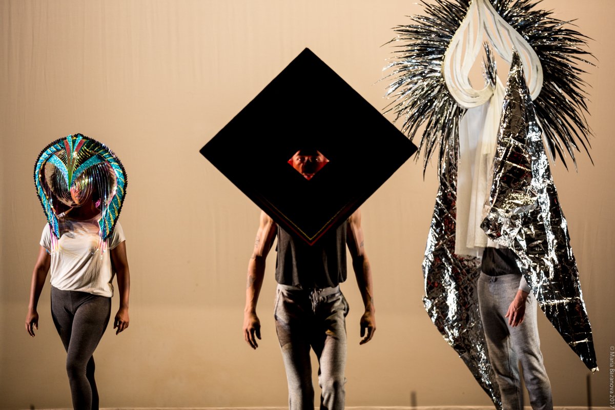 Three performers walk forward wearing various oversized masks (one a geometric diamond, the other two curvy, feathery, and fantastic). Photo by Maria Baranova.