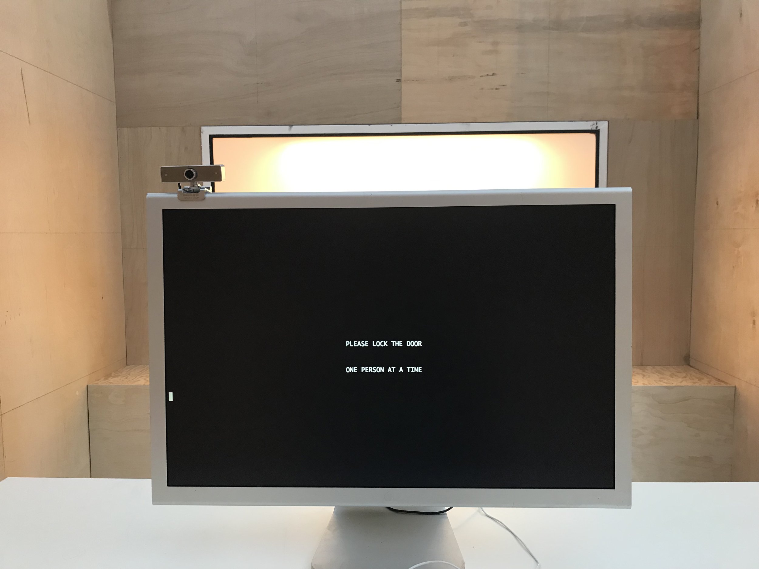 A computer screen display with the words “Please Lock The Door / One Person At A Time” inside the installation of a room made of wood panels. 