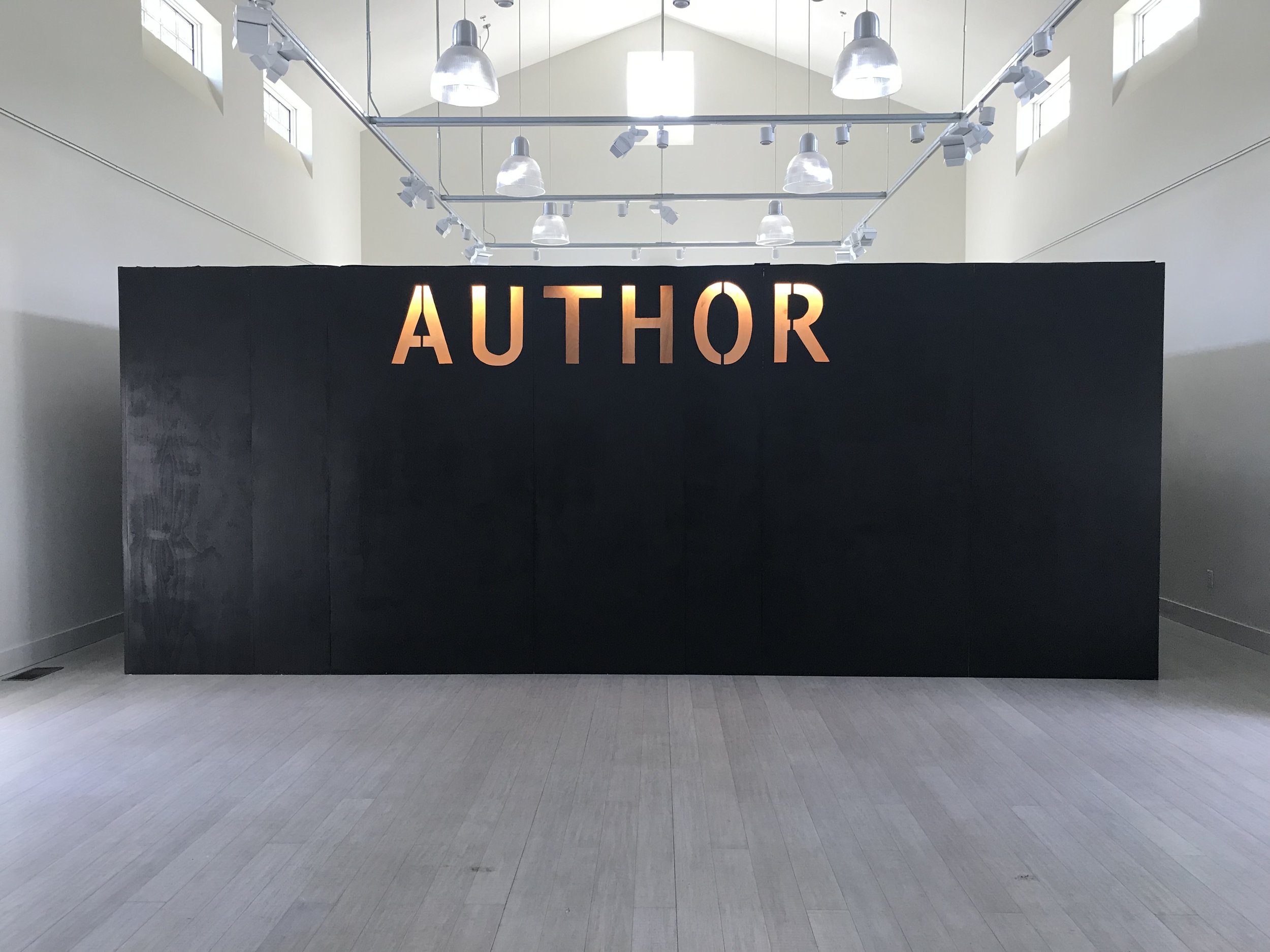 An exterior view of the installation – a simple black building with letters of “Author” carved into the side stands inside a white gallery space with vaulted ceilings.