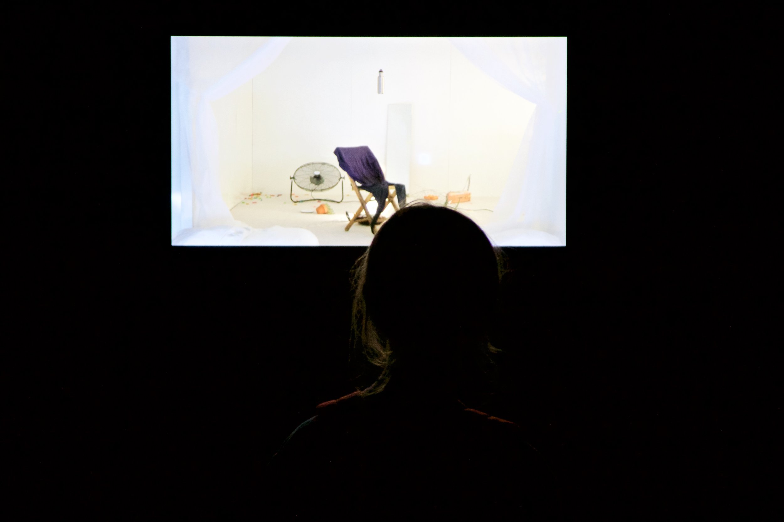 A person looks at a projected video (a chair and fan in white space framed by white curtains). Photo by Simon Courchel.