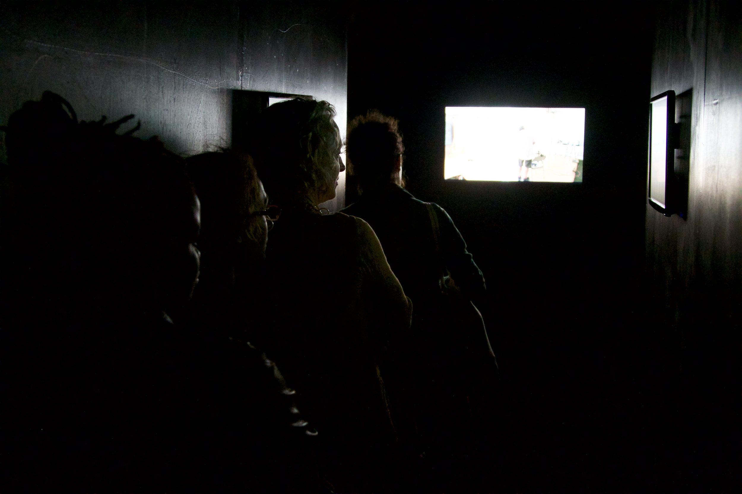 A line of people lean on a wall viewing various screens mounted on the walls of a dark, narrow hallway. Photo by Simon Courchel.