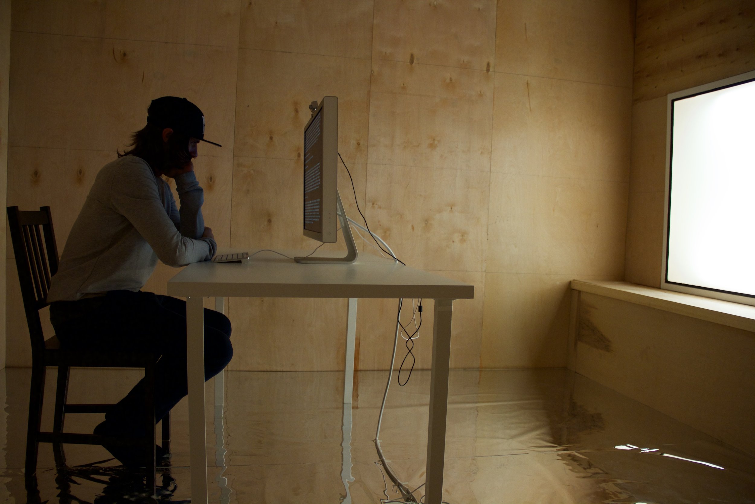 A person sits at a desk in front of a computer display reading text inside the installation of a room made of wood panels. Photo by Simon Courchel.