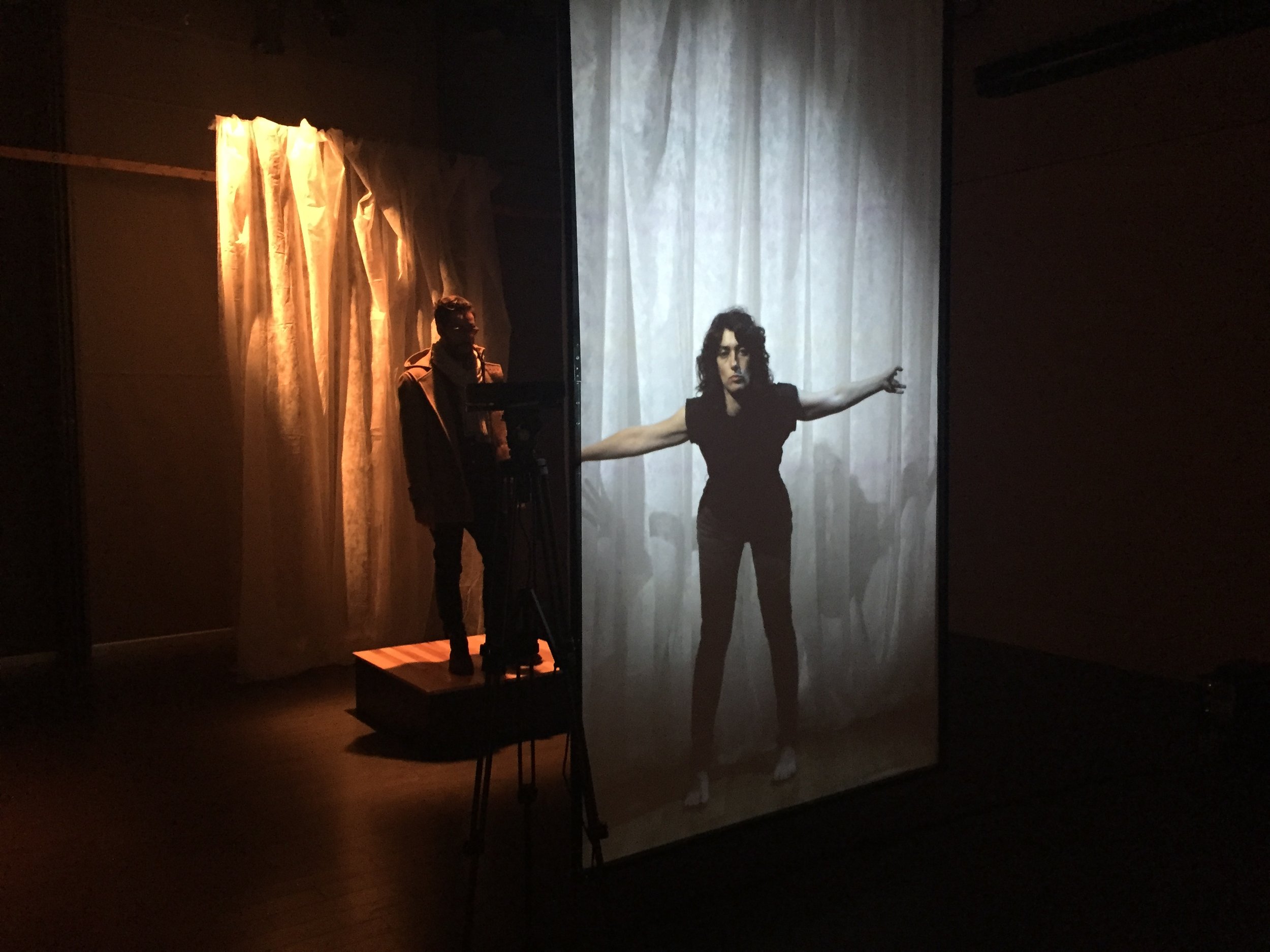 A person stands on a platform and interacts with a video (a performer gesturing in front of a white curtain.).
