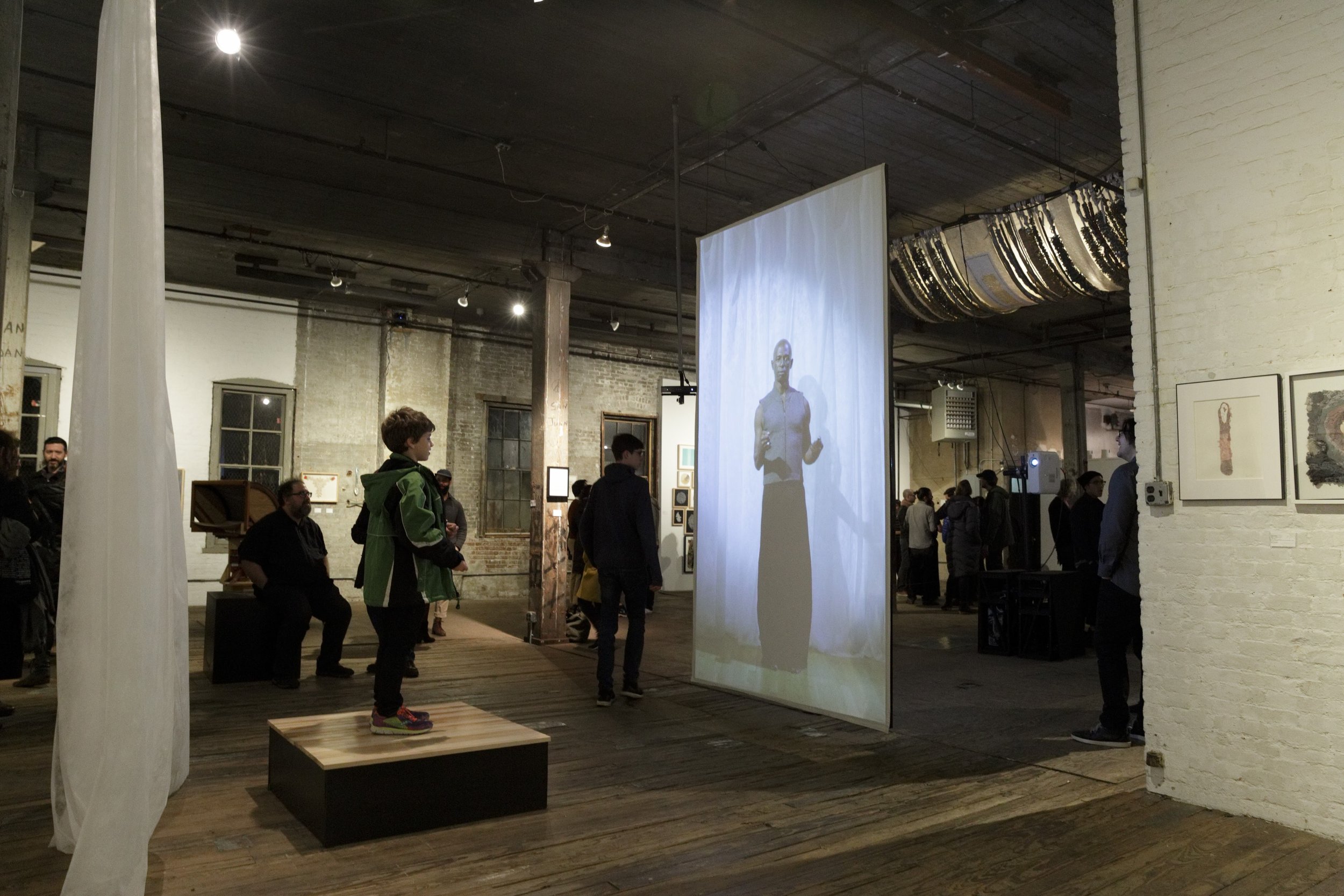 A child stands on a platform and interacts with a video (a performer gesturing in front of a white curtain) during a gallery opening event. 