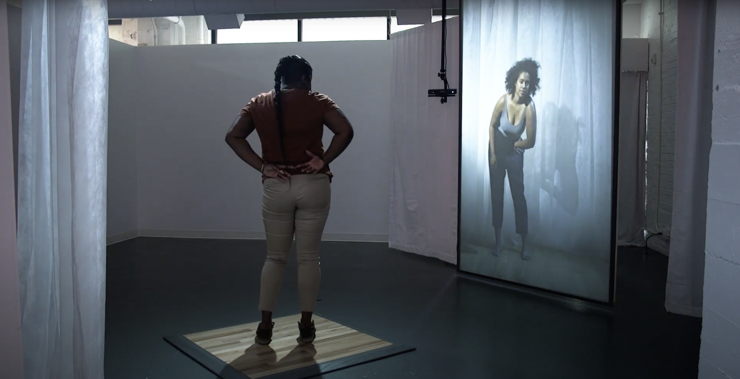 A person stands on a platform and interacts with a video (a performer gesturing in front of a white curtain) inside The Wndr Museum.