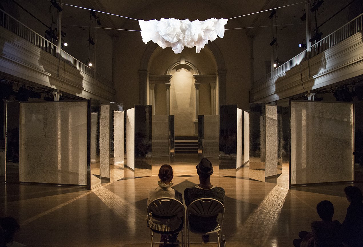 Two audience members sit in chairs viewing the opening stage set: a half circle of reflective panels below a suspended fluffy cloth cloud. Photo by Jose Espaillat.