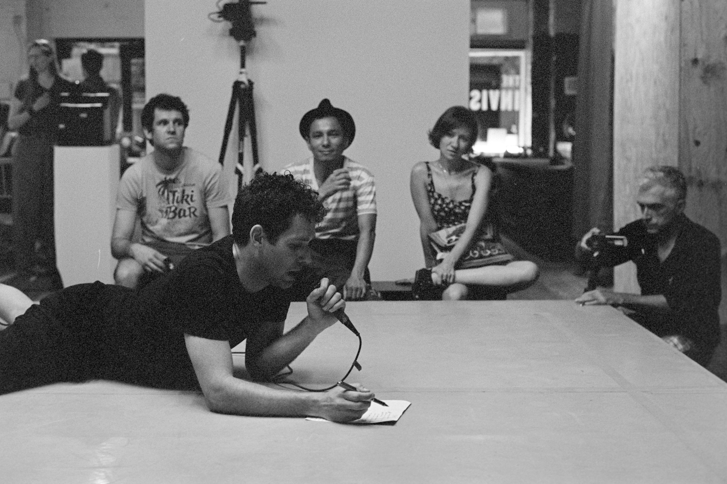 A performer lays on a platform speaking into a mic viewed by three people on a bench and a videographer. Photo by David Bivins.
