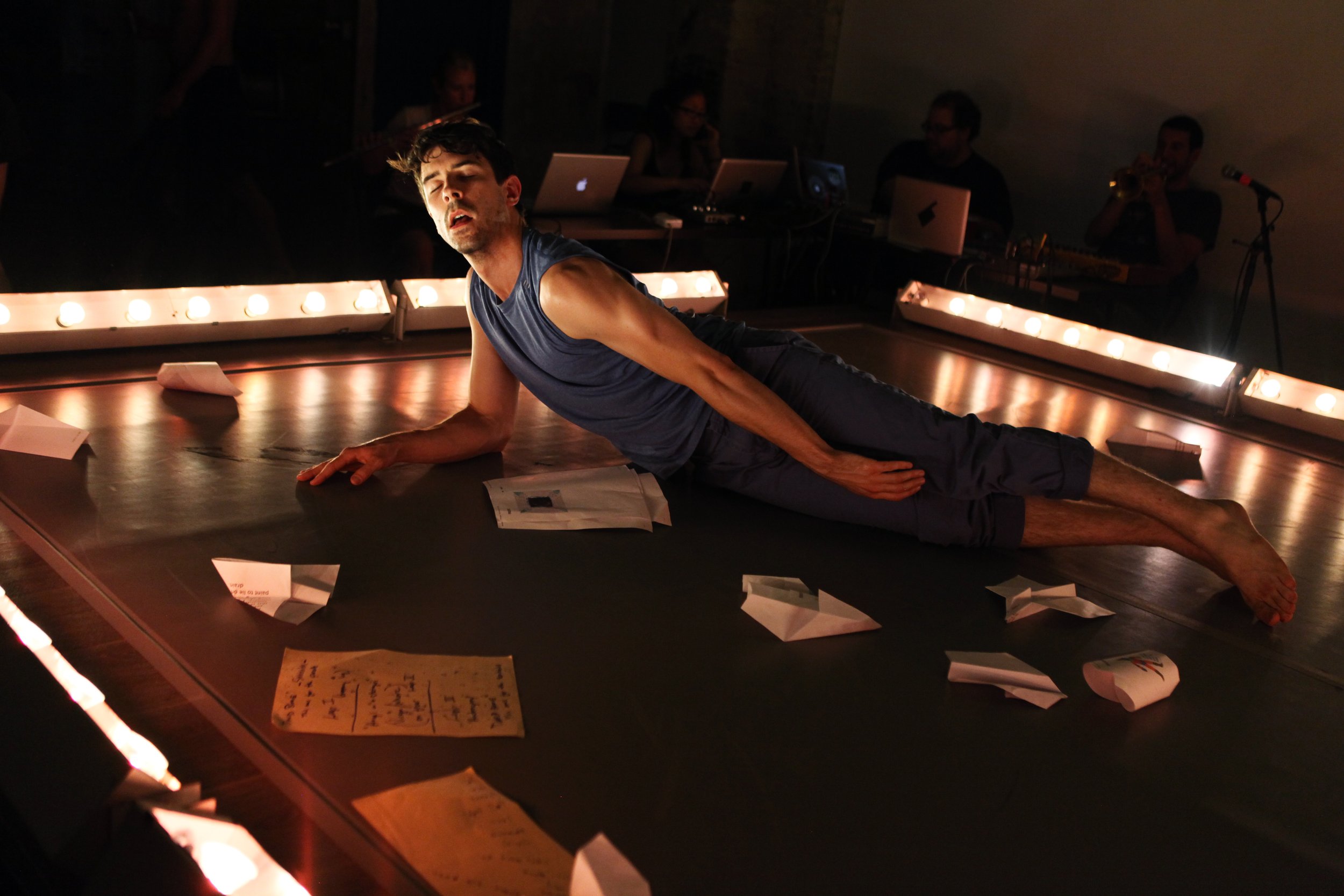 A performer extends his body laying on his side among many crumpled pages on a stage bordered by single rows of light boxes. Photo by Julieta Cervantes