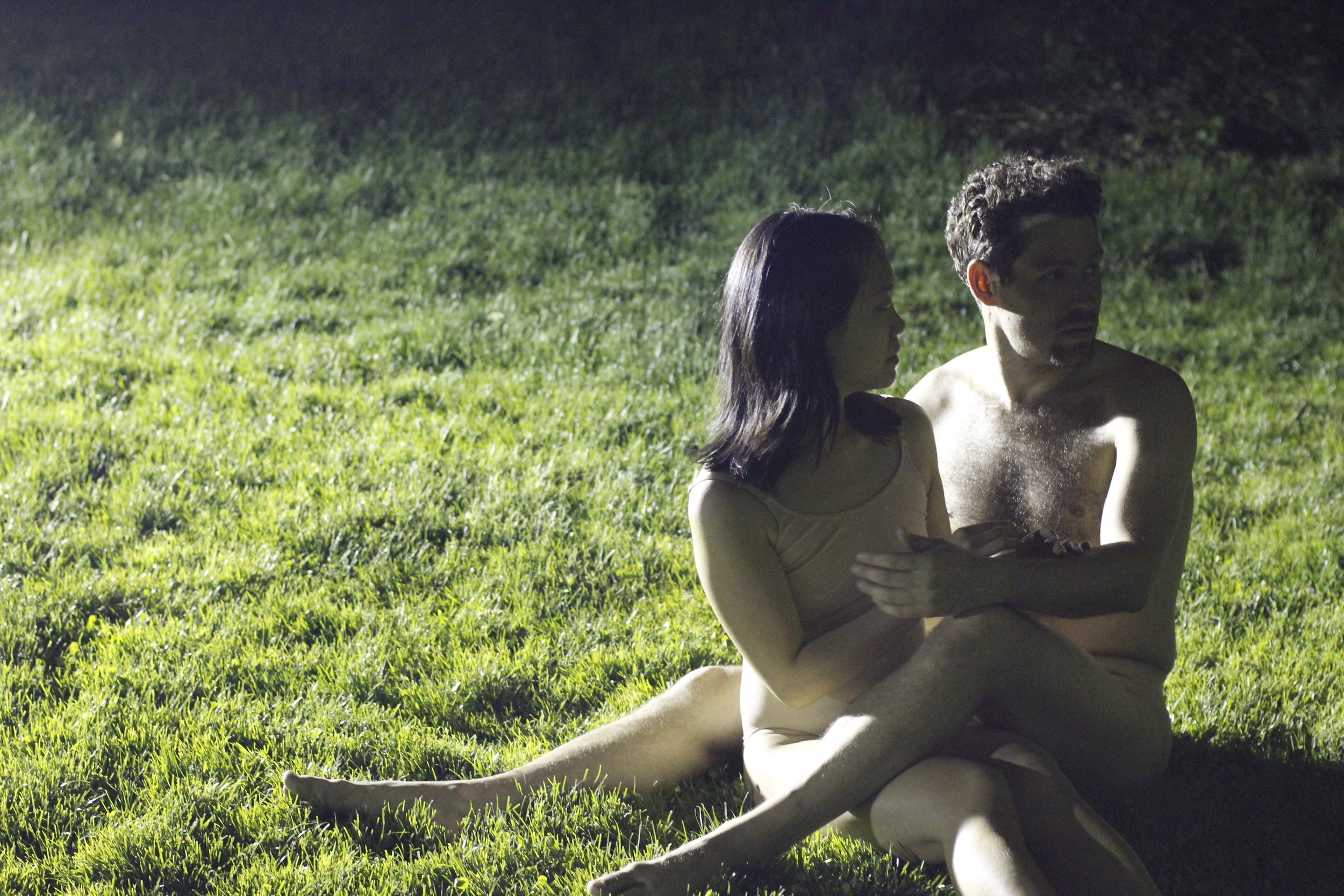 Two performers sit together, one's legs wrapped around the other, in a stream of light on green grass. Photo by Kevin Kwan.