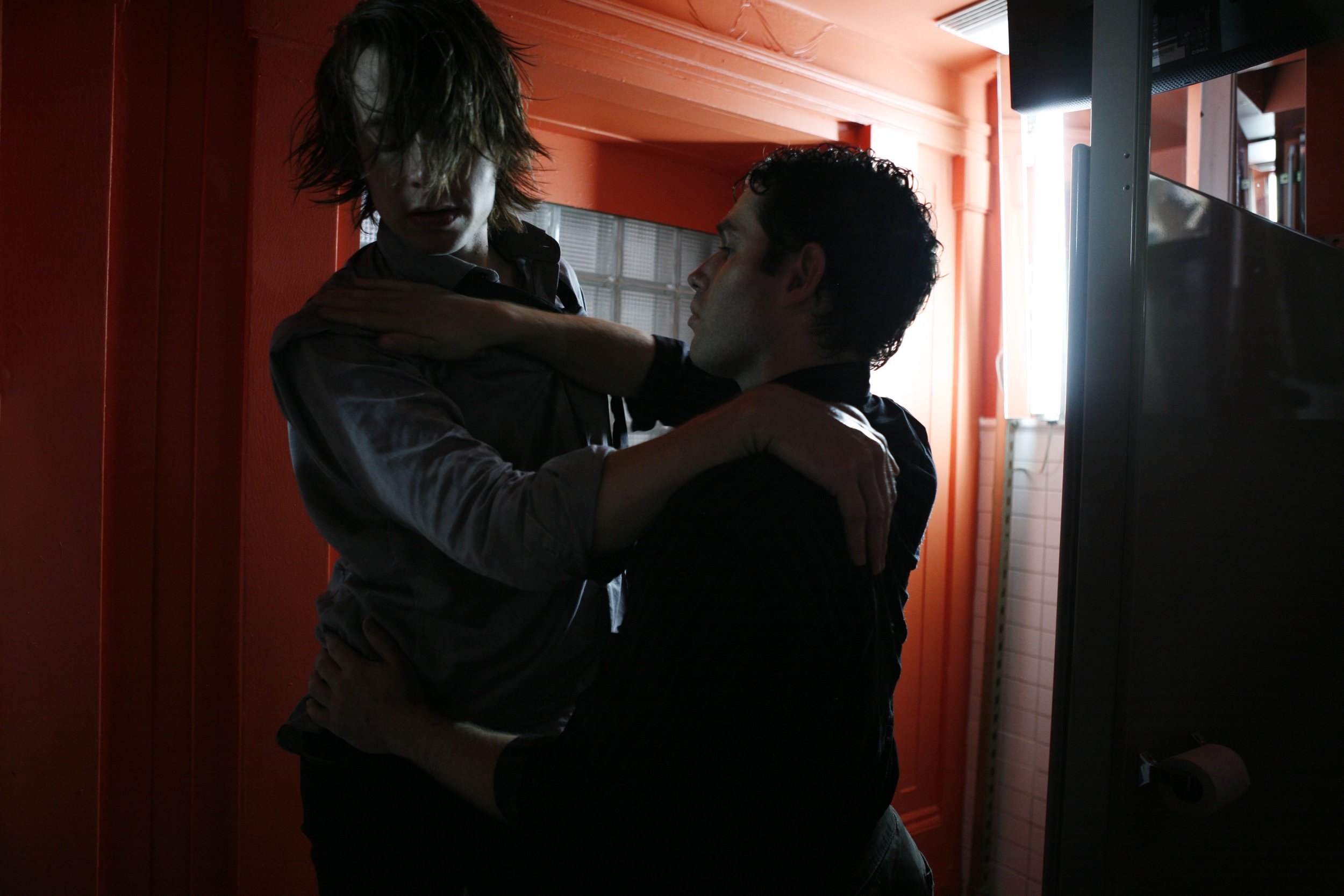 Two performers hold on to one another in the corner of a public restroom near a stall. Photo by Julieta Cervantes.