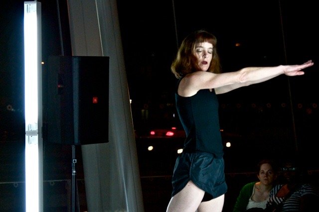 A performer in black shorts steps forward (arms pointed straight in front of her) lit by a column of fluorescent light. Photo by Simon Courchel. 