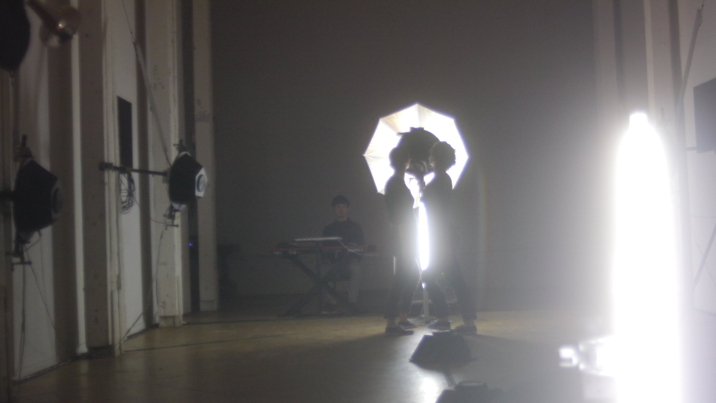 Two performers stand in front of a hemisphere speaker backlit by an umbrella light. A keyboardist sits nearby in a narrow hall. Photo by John Preston Gomez.