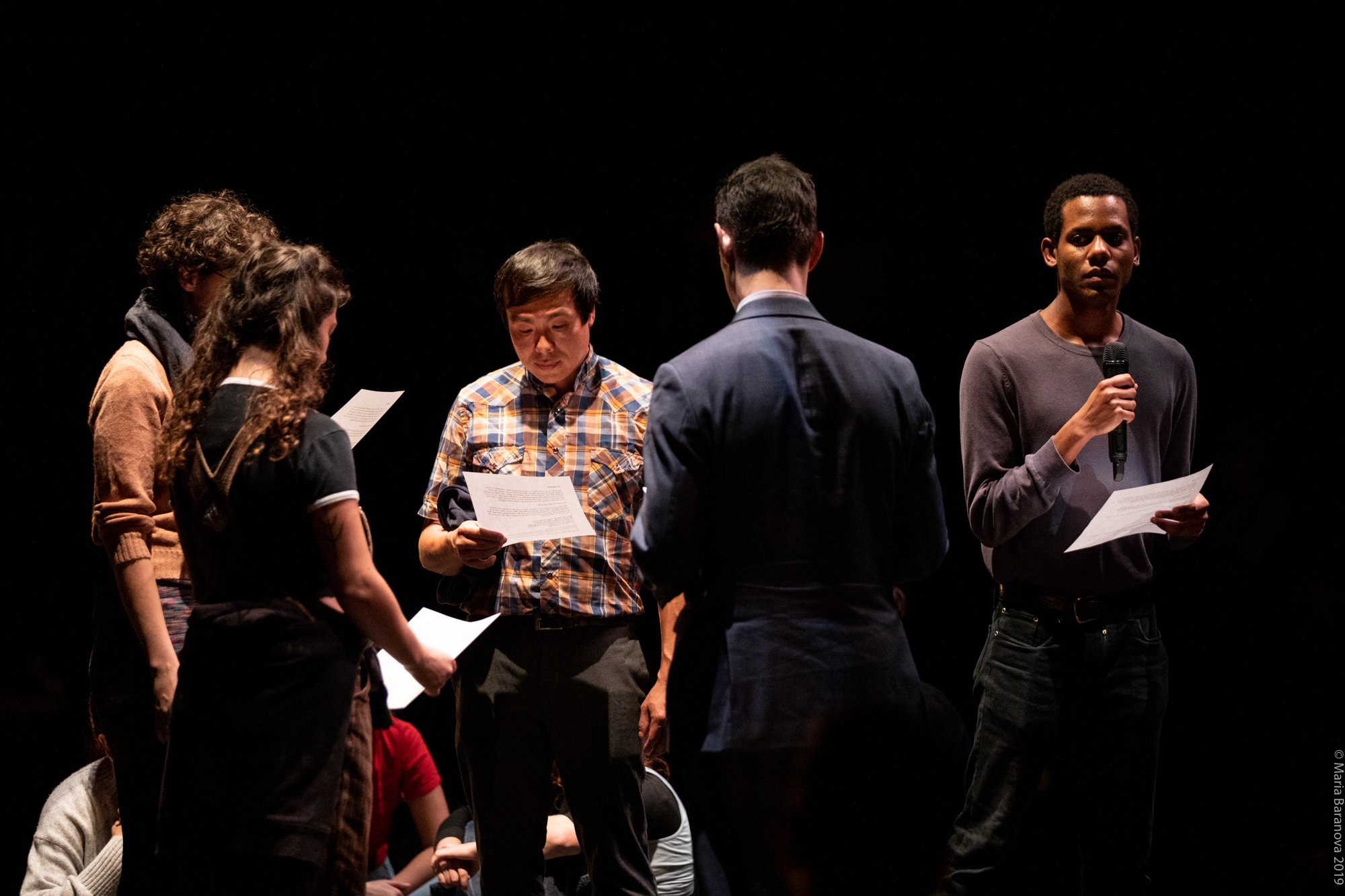 A group of people holding white paper scripts and engaged in an action together stand in a theater space. Photo by Maria Baranova.
