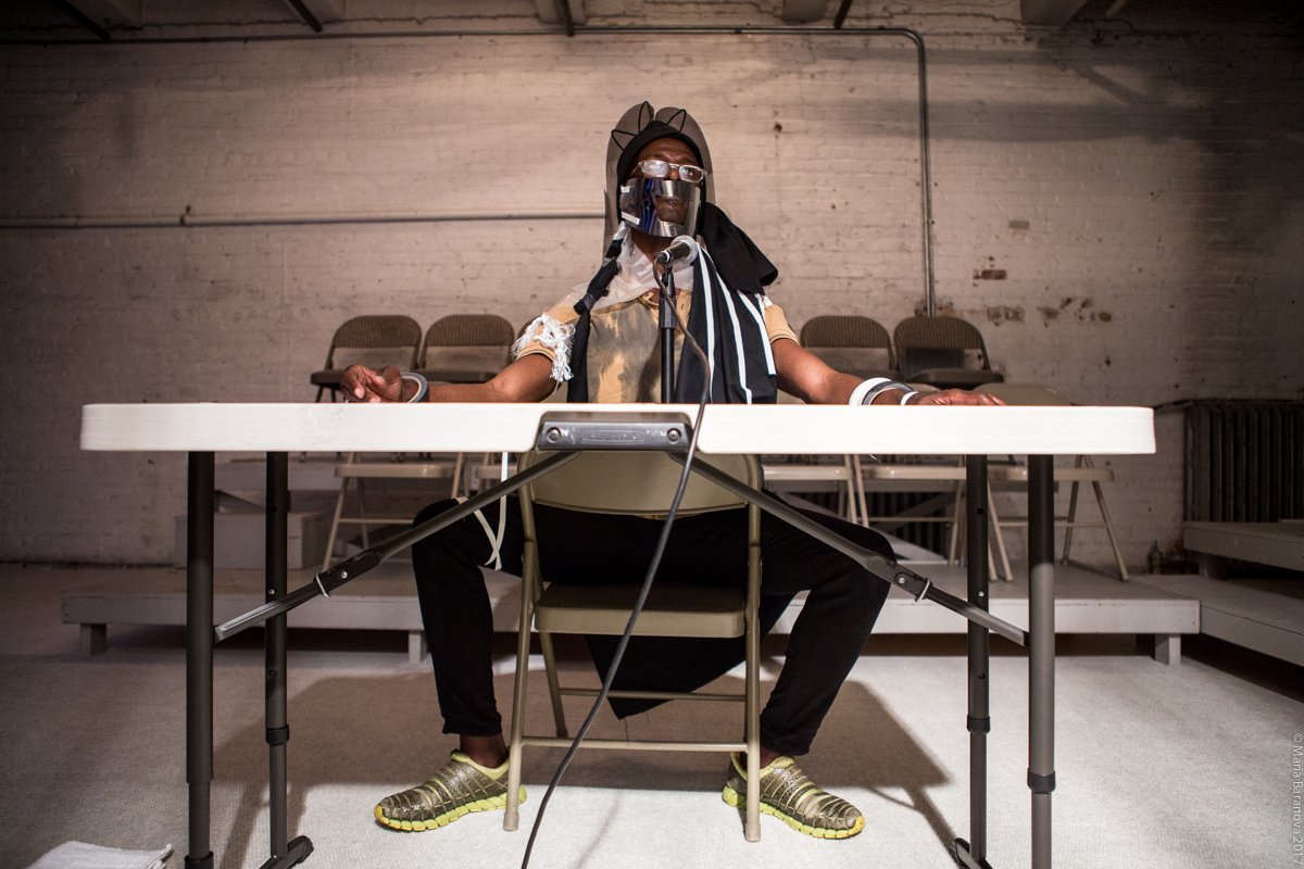 A performer in glasses, his torso and head wrapped in fabrics and tape, sits behind a table and a mic in a performance space. Photo by Maria Baranova. 