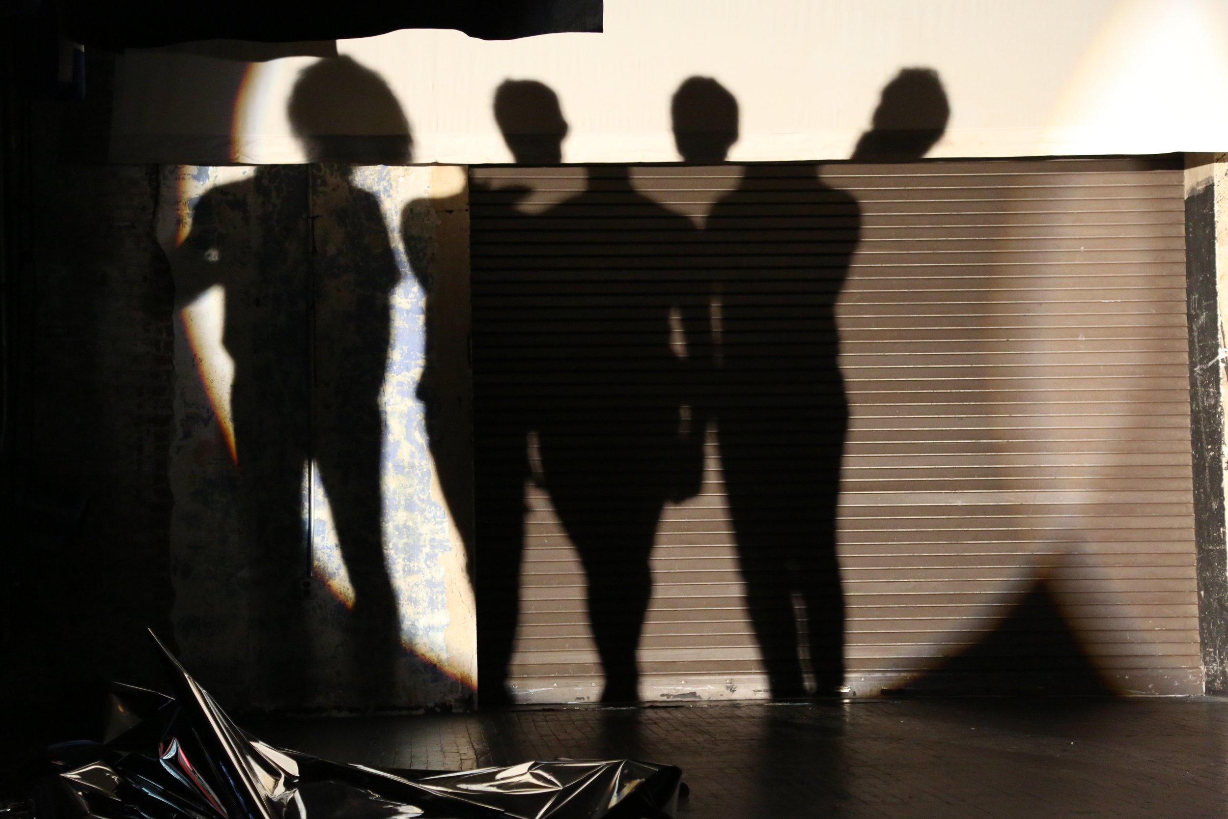 The shadows of four performers standing together are cast on the back wall of a stage in a large spotlight. Photo by Brian Rogers.