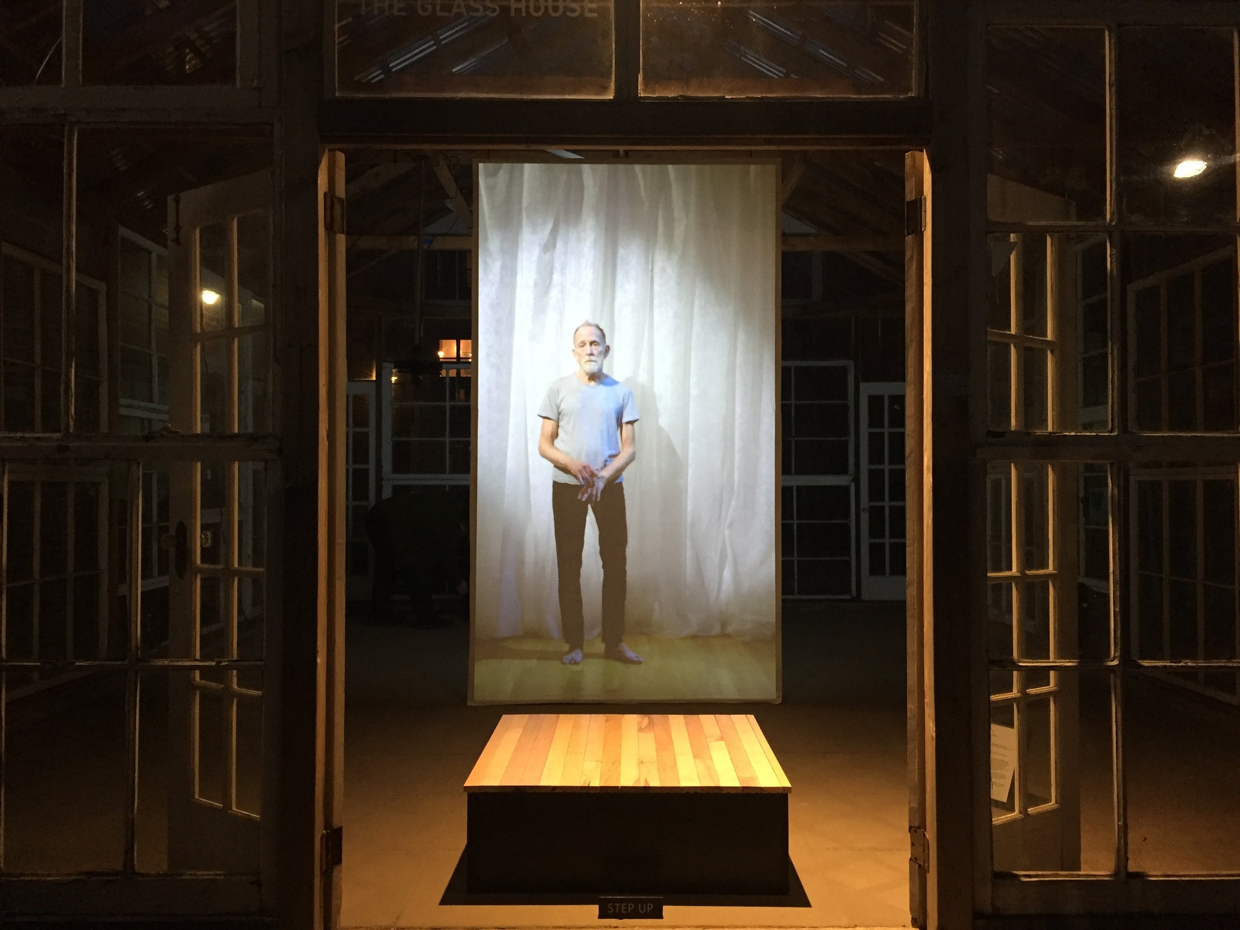 A projection of a performer standing in front of a white curtain hangs by a small square platform inside a glass house at night.