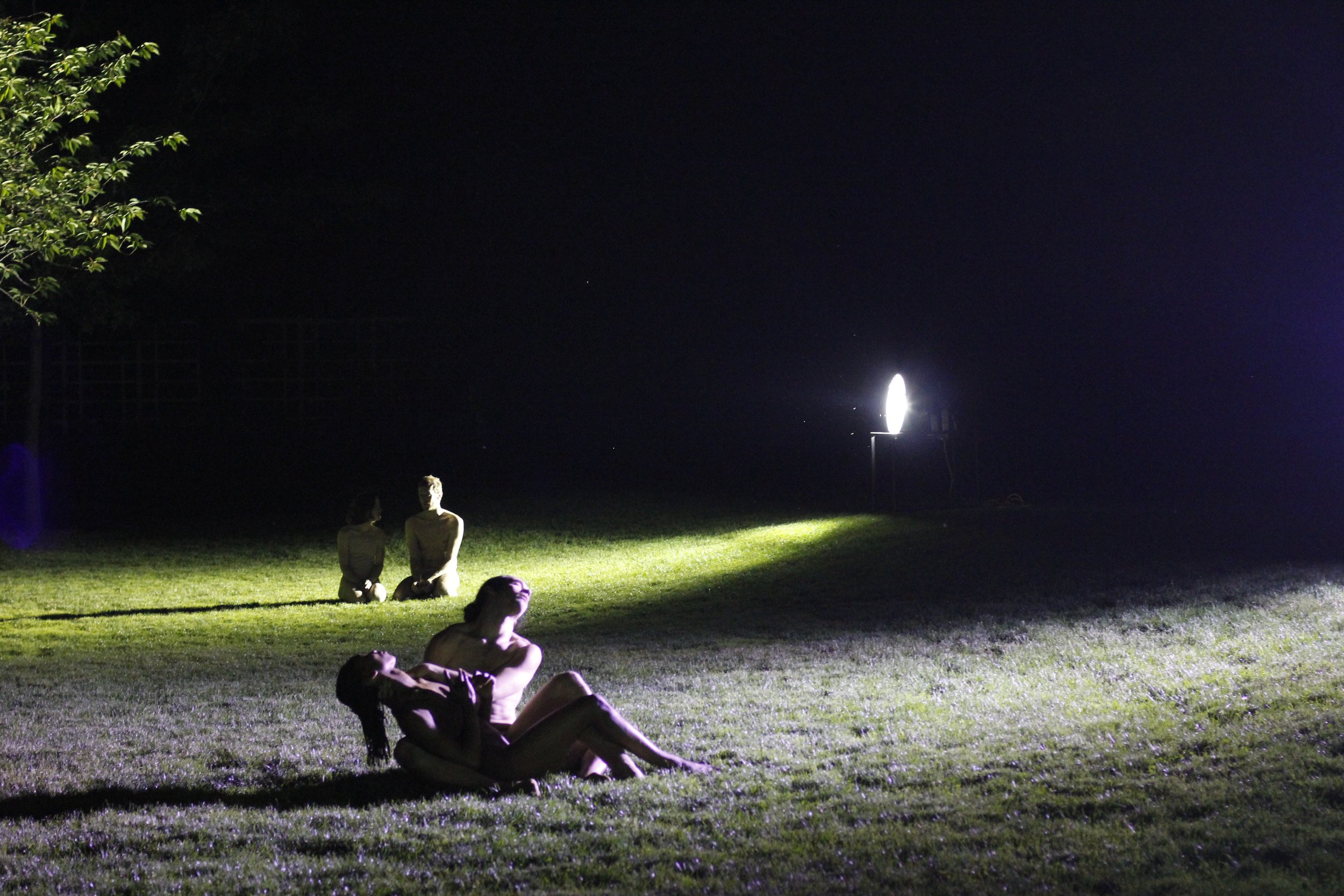 Two groups of two performers sitting together in streams of light on green grass. Some leafy branches of a tree overhead. Photo by Kevin Kwan.