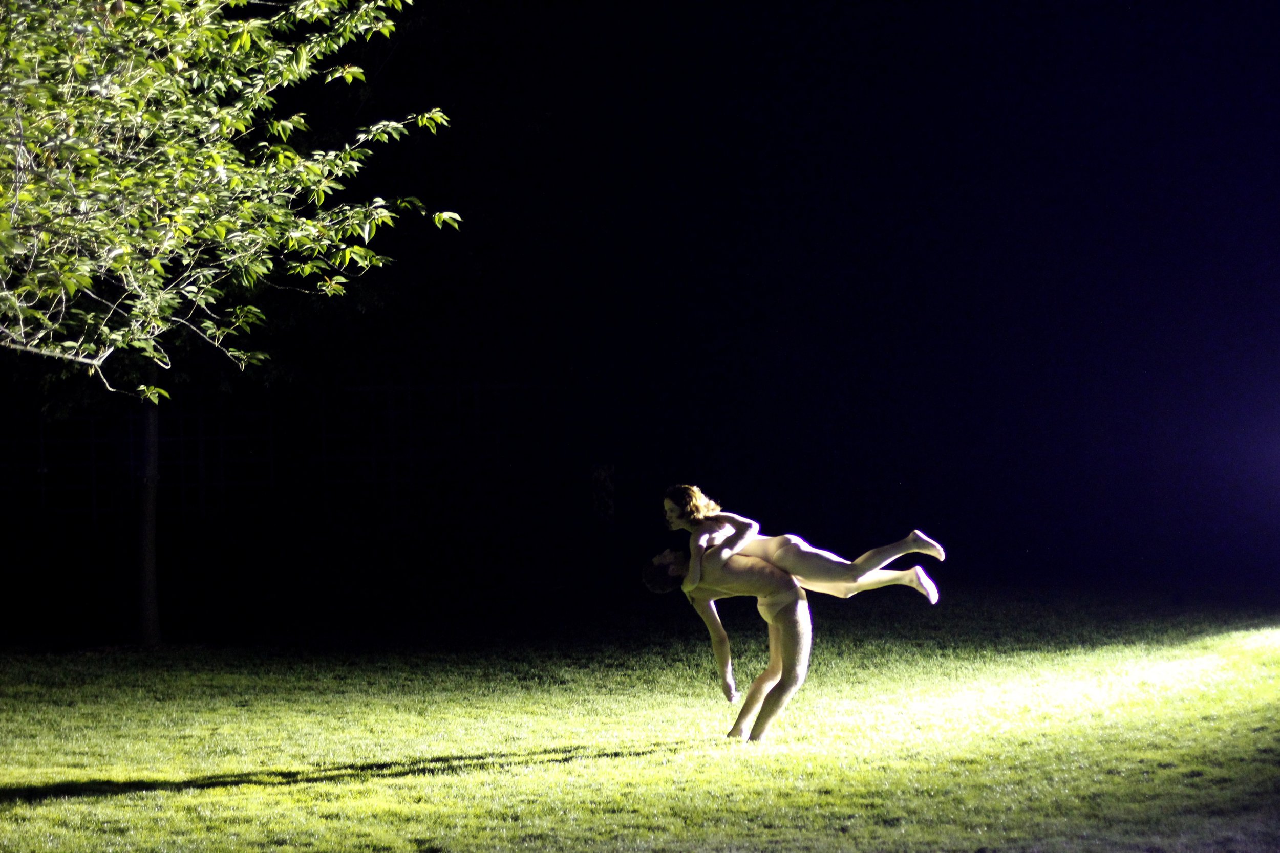 Two performers (one lifted off the ground by the other) in a stream of light on green grass. Photo by Kevin Kwan.