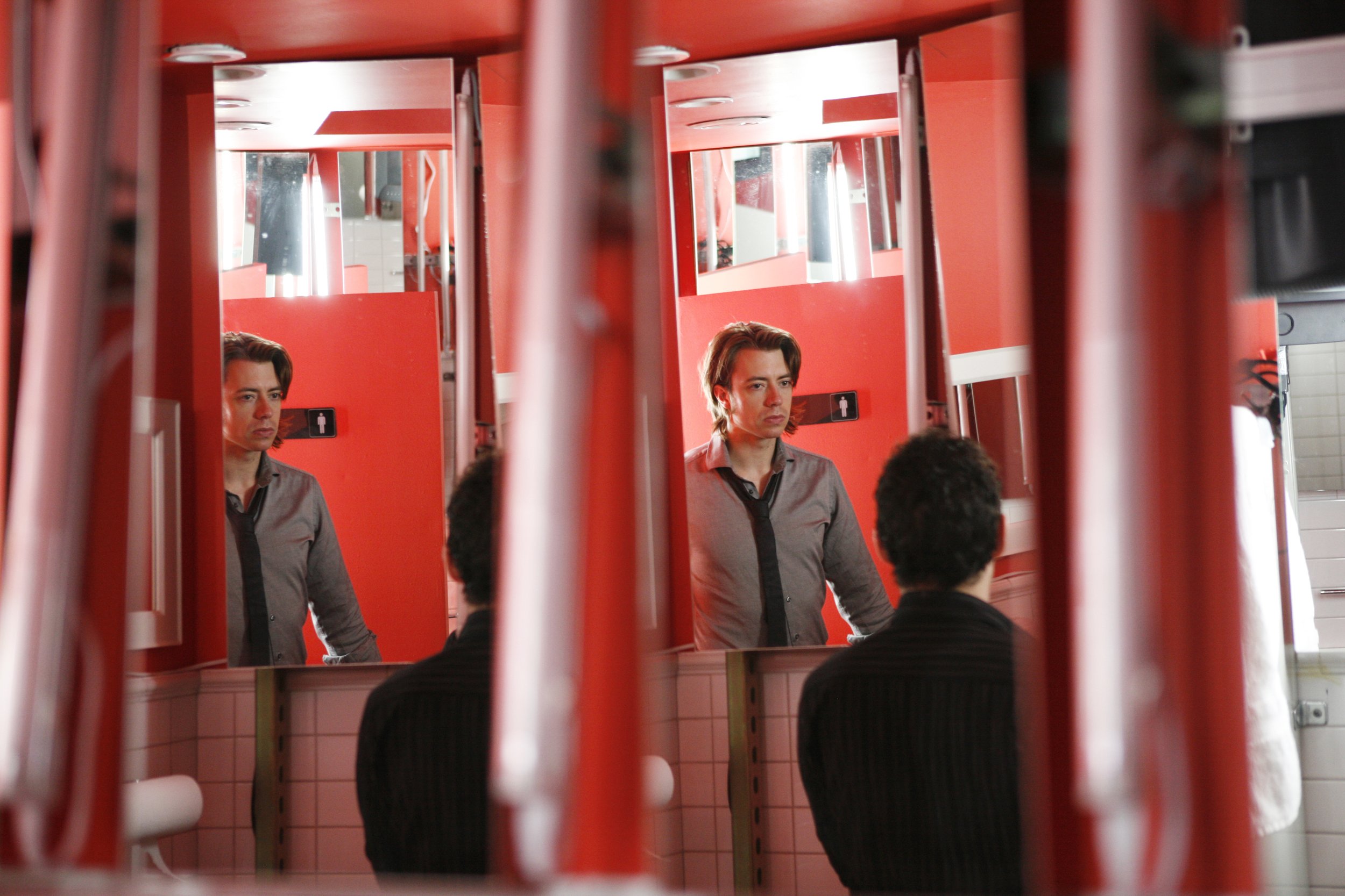 The duplicated reflections of a performer looking at the reflection of another performer in a row of mirrors in a public restroom. Photo by Julieta Cervantes.