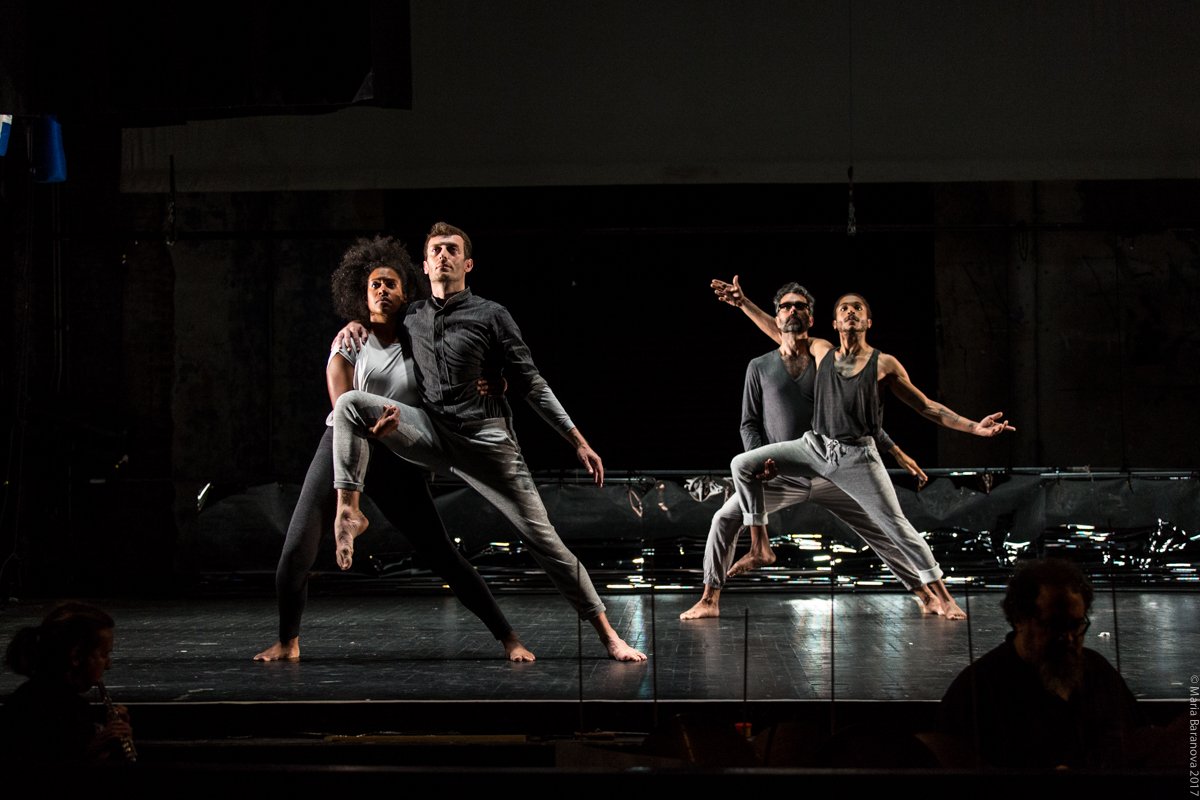 Four performers lunging to the side, looking out performatively, and holding each other in groups of two on a stage. Photo by Maria Baranova.